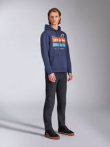 Alpinestars Harken Hoodie en azul marino, sudadera con capucha usada por un modelo masculino con pantalones vaqueros oscuros y zapatillas negras, con un gran gráfico en el pecho con detalles en naranja y azul claro, logotipo Alpinestars y bolsillo canguro