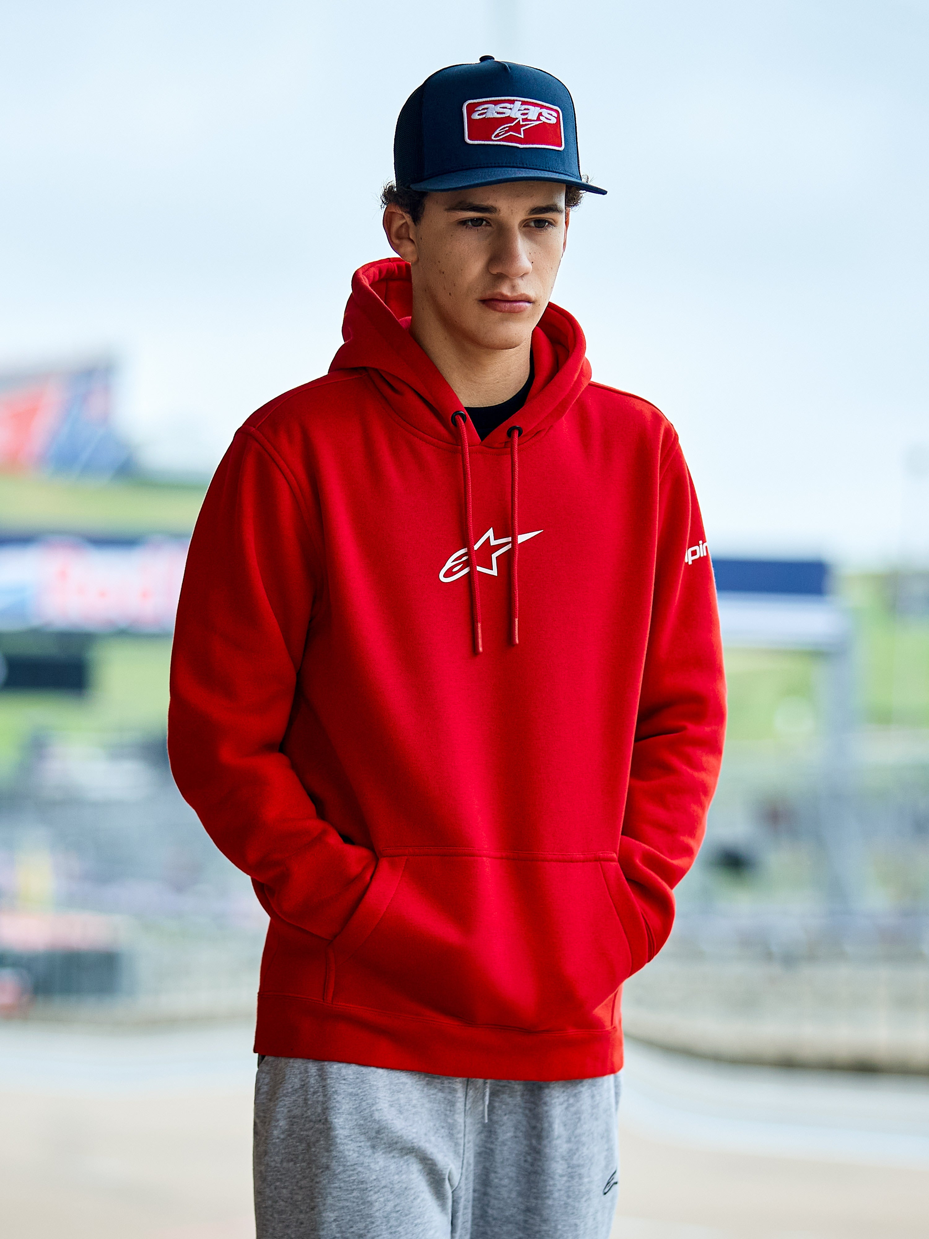 Alpinestars Frontal Hoodie, sudadera roja de forro polar con logotipo Astars blanco en el pecho, bolsillo canguro y capucha con cordón, usada por un hombre con gorra azul, estilo casual motorsport streetwear