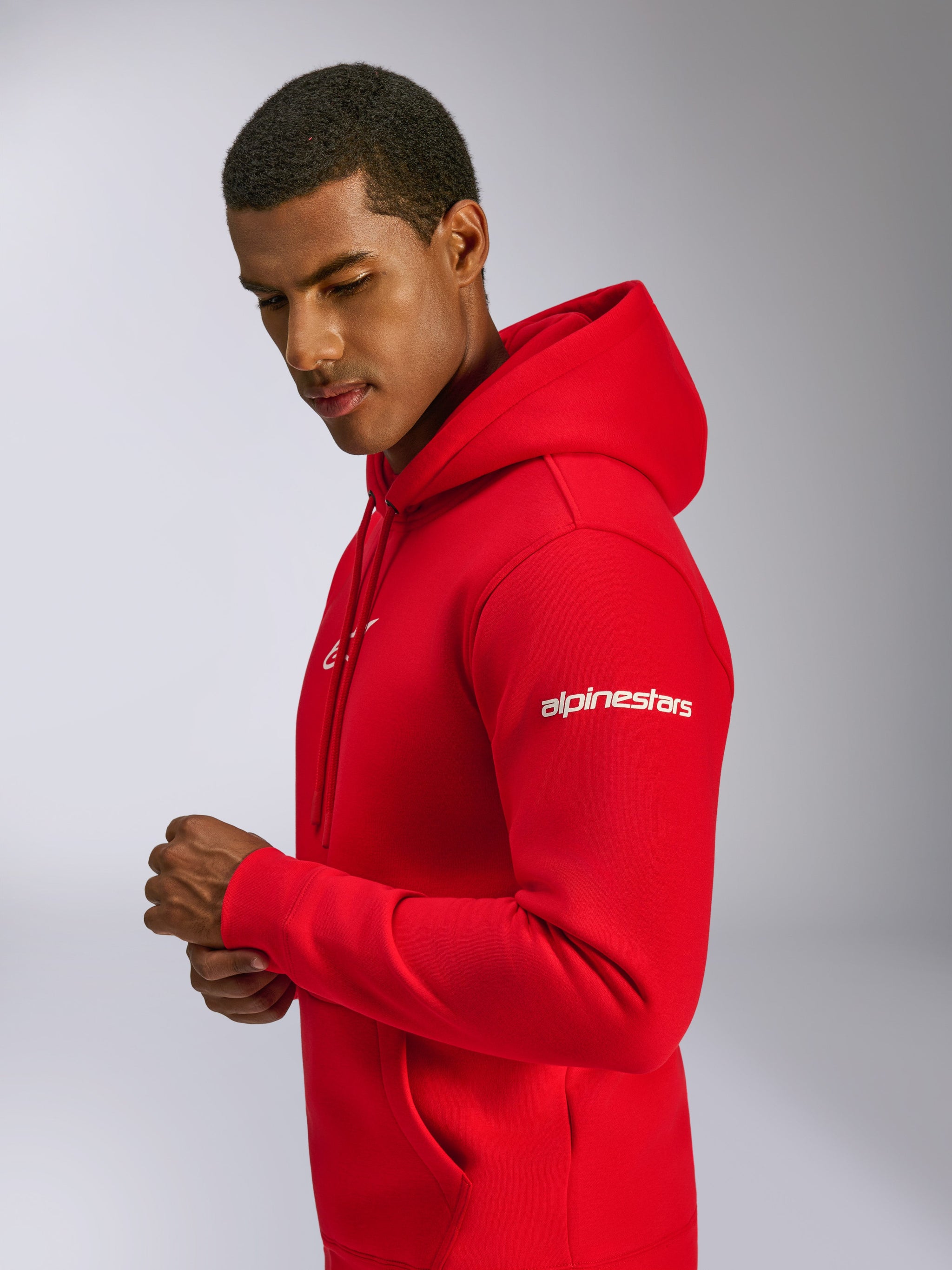 Sudadera con capucha Alpinestars Frontal, roja, sudadera de forro polar tipo pullover lucida por un modelo, con el logotipo Alpinestars blanco en la manga, logotipo frontal, bolsillo canguro y capucha con cordón, diseño casual inspirado en el automovilismo