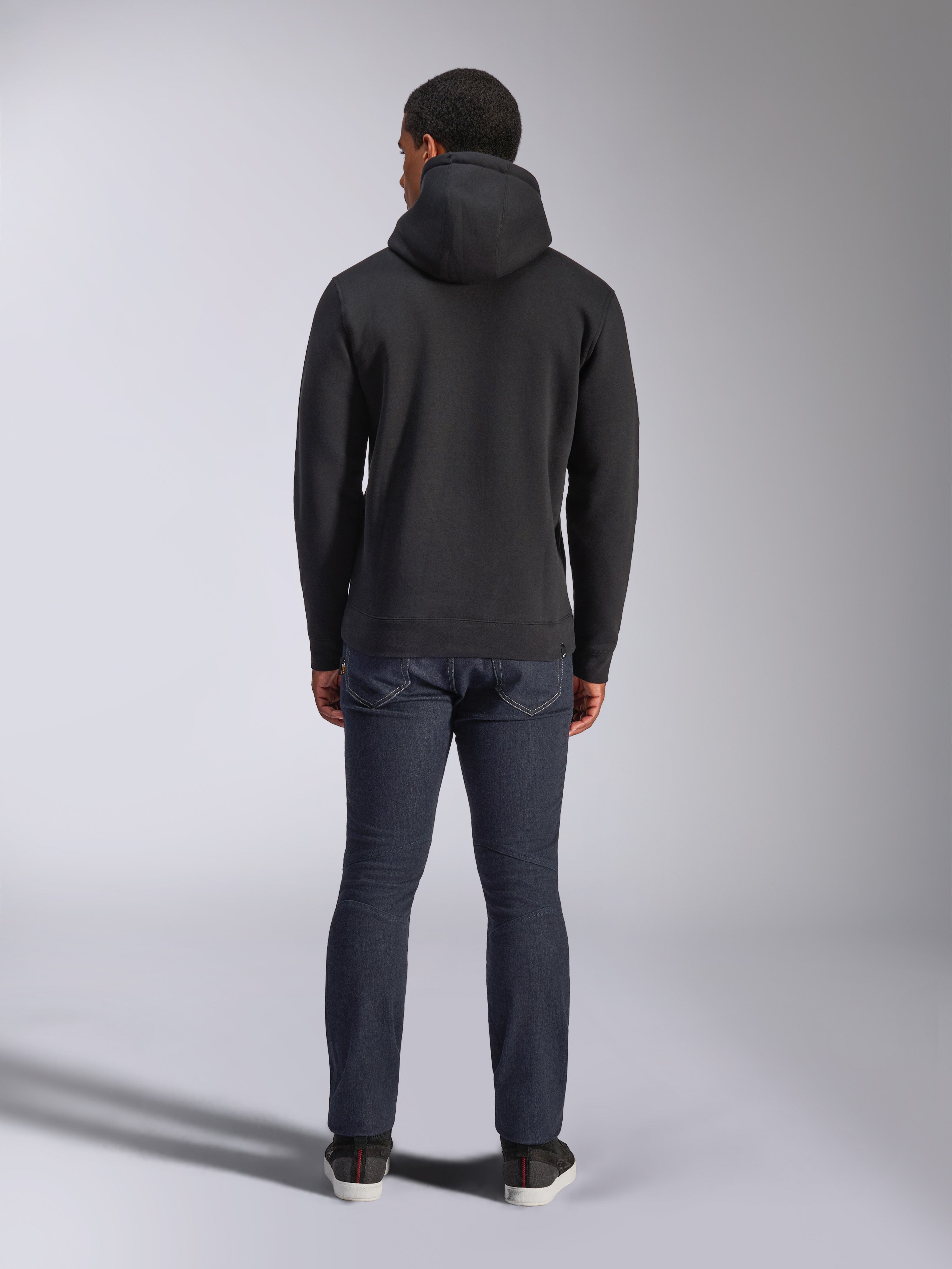 Sudadera con capucha Alpinestars Blaze V3, estilo urbano casual, negro, vista posterior de sudadera de felpa con capucha, combinada con vaqueros denim azul oscuro y zapatos negros, diseño limpio inspirado en el automovilismo