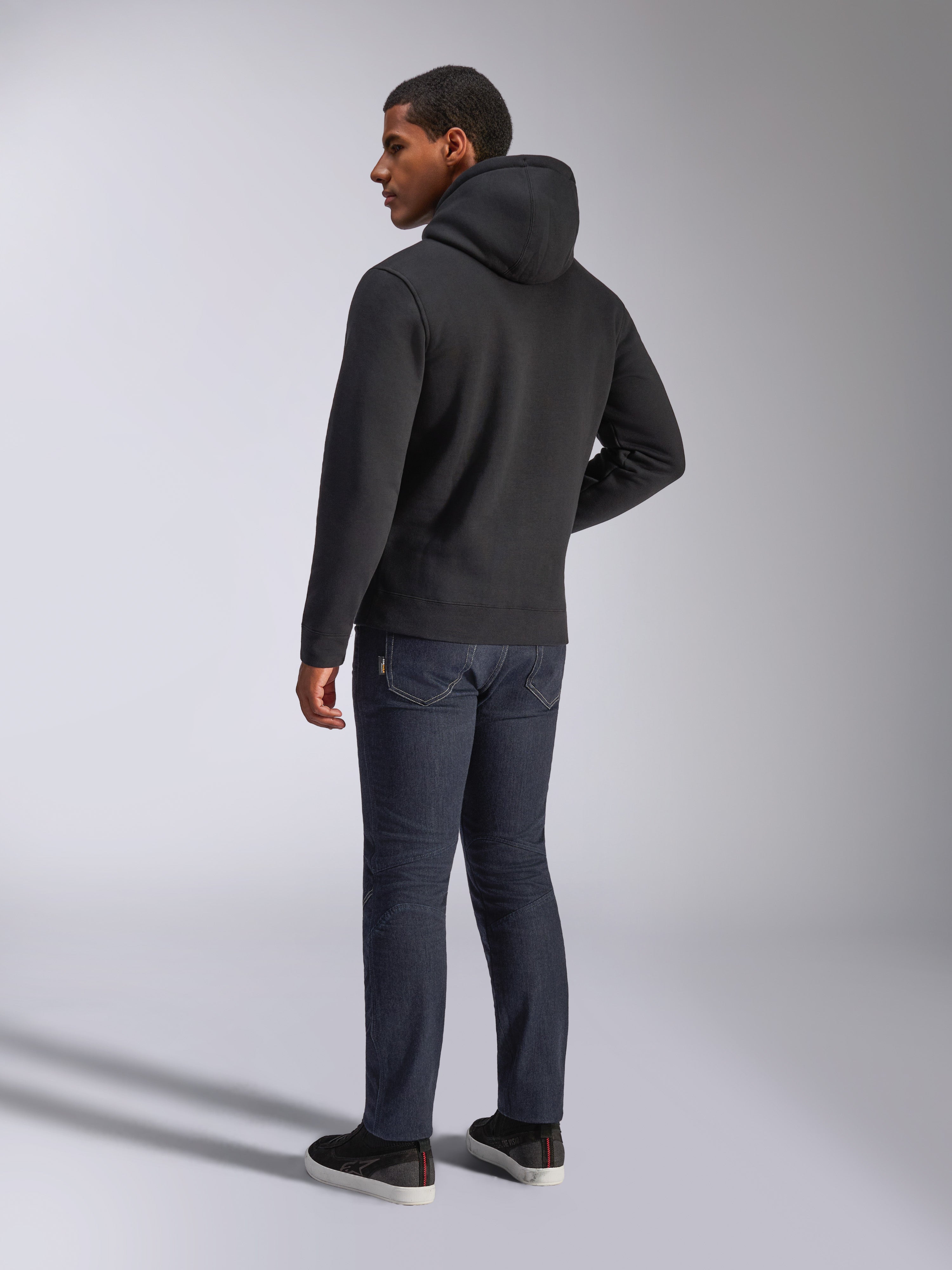 Alpinestars Blaze V3 Hoodie, vista posterior de un hombre con la sudadera negra, combinada con vaqueros oscuros y zapatillas negras, ropa urbana casual de inspiración motorsport