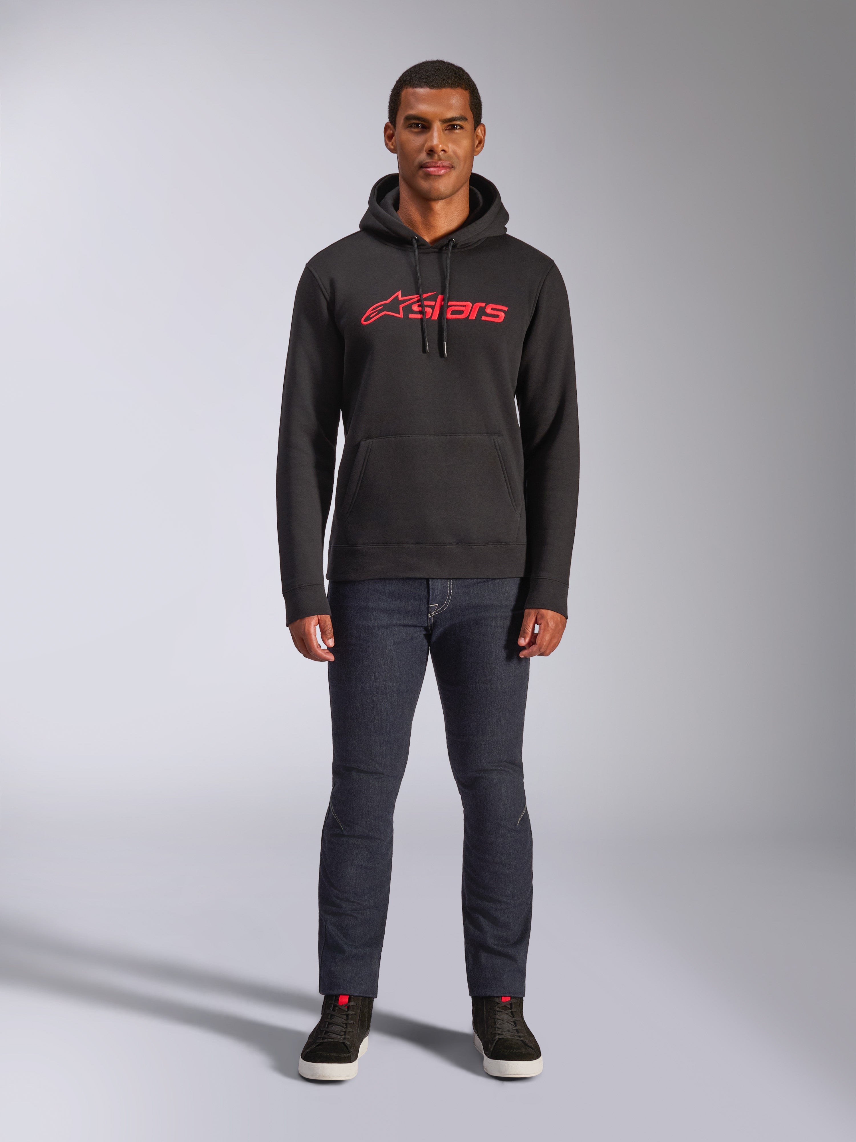 Sudadera Alpinestars Blaze V3 Hoodie, ropa urbana casual, negra con el logotipo Astars en rojo, vista frontal, modelo luciendo pantalones vaqueros oscuros y zapatillas negras, diseño de forro polar moderno inspirado en el automovilismo