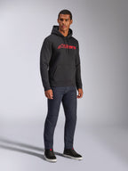 Sudadera con capucha Alpinestars Blaze V3, sudadera de felpa negra con el logotipo rojo de Alpinestars en el pecho, lucida por un modelo masculino con vaqueros de denim oscuro y zapatillas negras, estilo streetwear casual inspirado en el motor