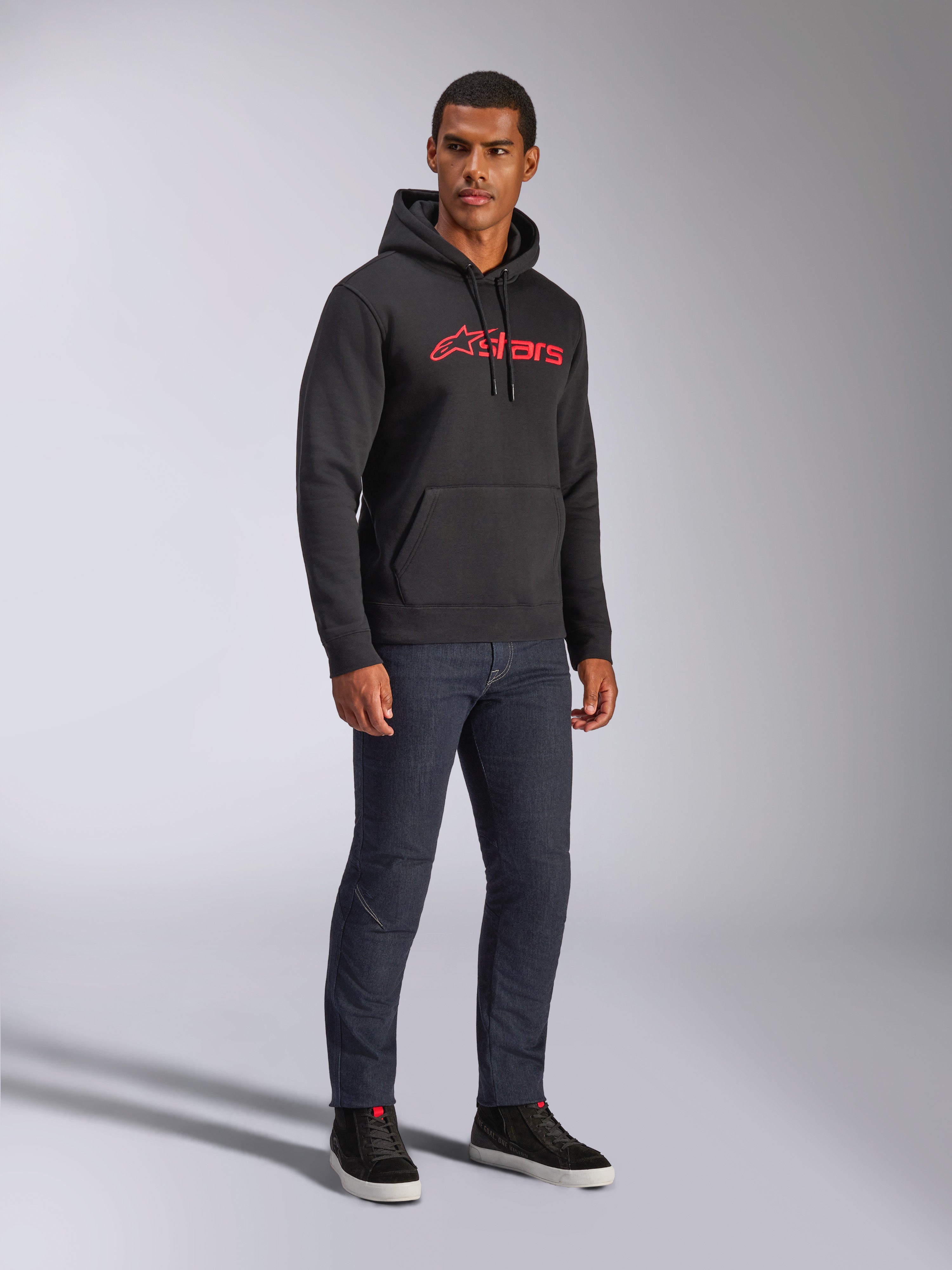 Sudadera con capucha Alpinestars Blaze V3, sudadera de felpa negra con el logotipo rojo de Alpinestars en el pecho, lucida por un modelo masculino con vaqueros de denim oscuro y zapatillas negras, estilo streetwear casual inspirado en el motor