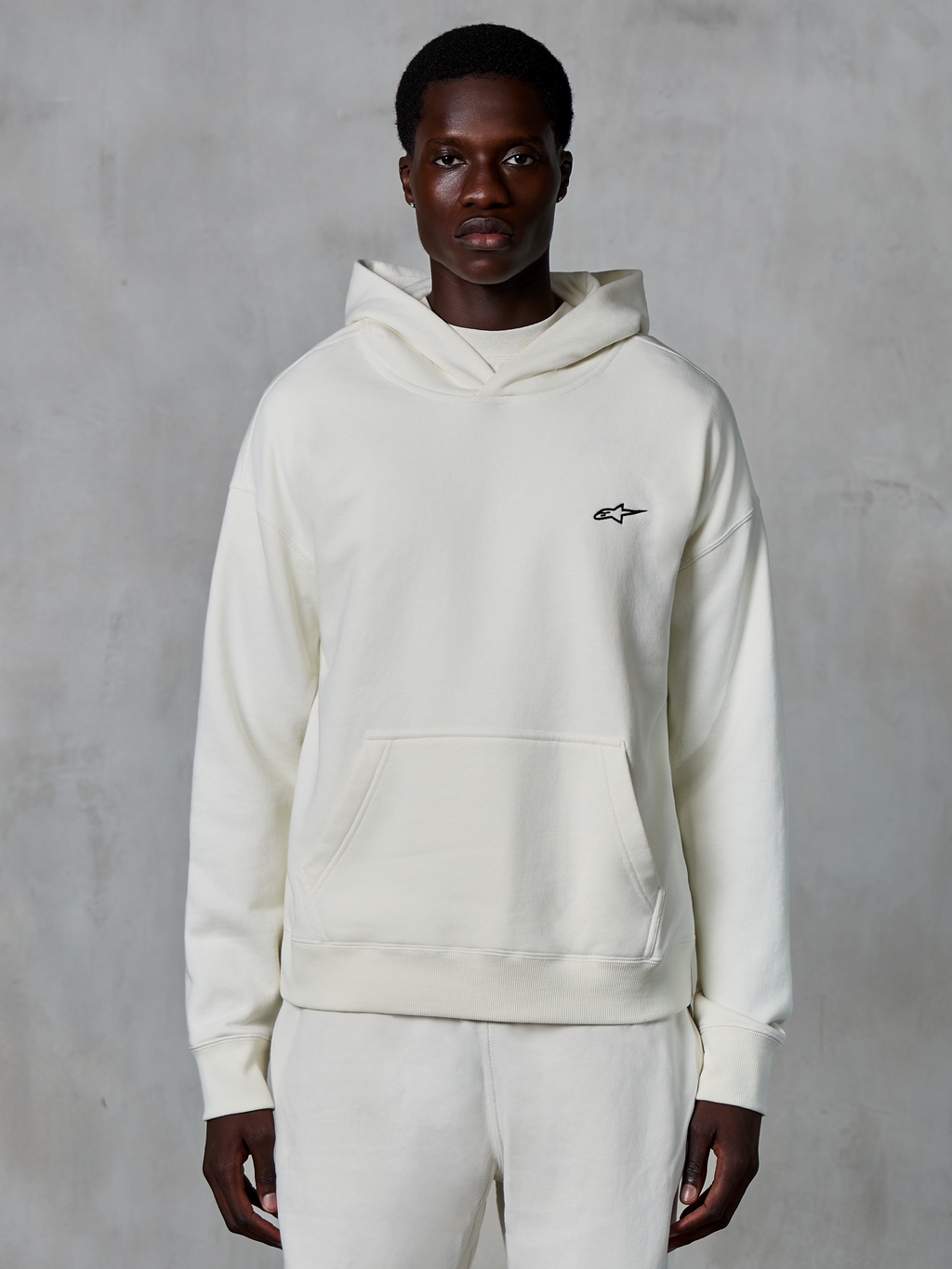 Alpinestars Essentials Ageless Hoodie, color blanco roto, usada por modelo masculino, pequeño logotipo negro en el pecho, bolsillo tipo canguro frontal, manga larga, ropa urbana casual inspirada en el automovilismo