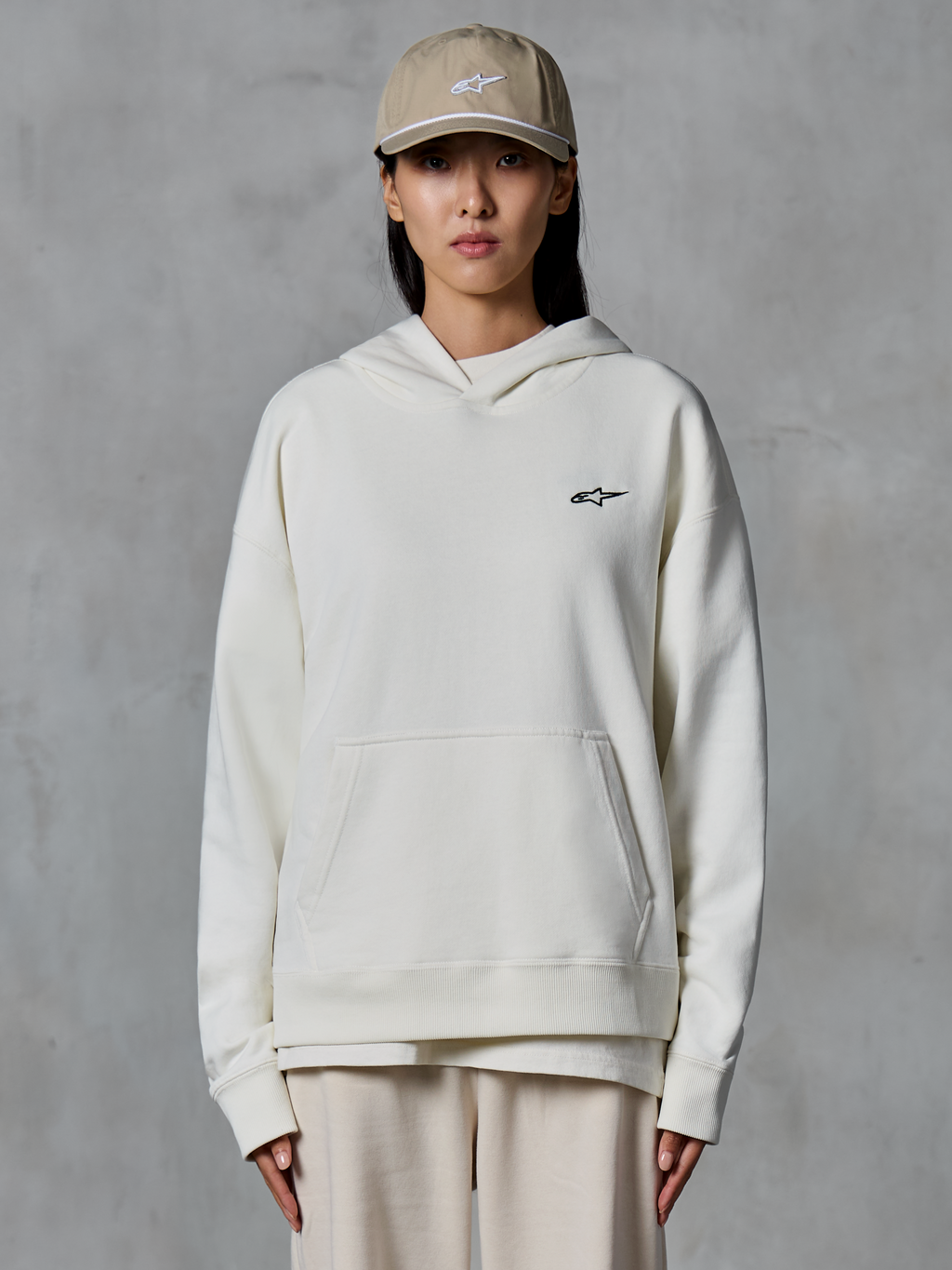 Alpinestars Essentials Ageless Hoodie, sudadera con capucha de forro polar en color blanco roto, modelo femenina con gorra color tostado y joggers crema a juego, pequeño logotipo negro de Alpinestars en el pecho, bolsillo de canguro delantero, ajuste relajado, estilo urbano casual inspirado en el motociclismo
