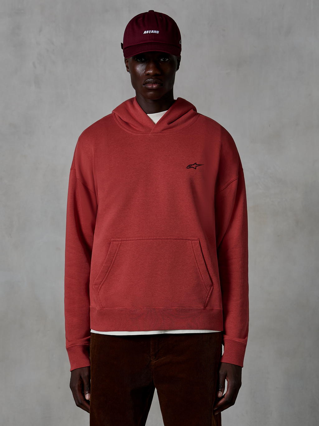 Sudadera con capucha Alpinestars Essentials Ageless, ropa urbana casual, color rojo Brick, con un pequeño logo de estrella negra en el pecho, bolsillo tipo canguro y acabado de lavado a la piedra, llevada por un modelo con gorra granate y pantalones de pana marrones