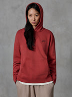 Alpinestars Essentials Ageless Hoodie, sudadera de color rojo ladrillo, usada por una modelo con la capucha puesta, con un pequeño logo de estrella bordado en negro en el pecho, bolsillo tipo canguro, ropa urbana casual de estilo de vida inspirada en el motociclismo