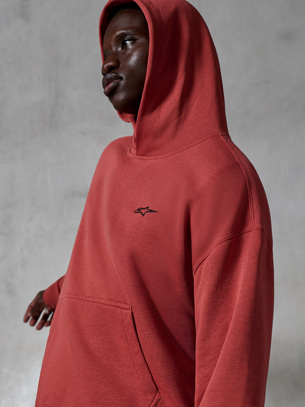 Sudadera con capucha Alpinestars Essentials Ageless, ropa deportiva casual, color rojo teja, lucida por un modelo con la capucha puesta, con un pequeño logotipo negro bordado, textura de forro polar suave, estilo urbano de corte relajado