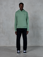Alpinestars Essentials Ageless Hoodie, Cactus green, small black chest logo, kangaroo pocket, casual lifestyle streetwear worn with black pants and black and white sneakers against a textured wall