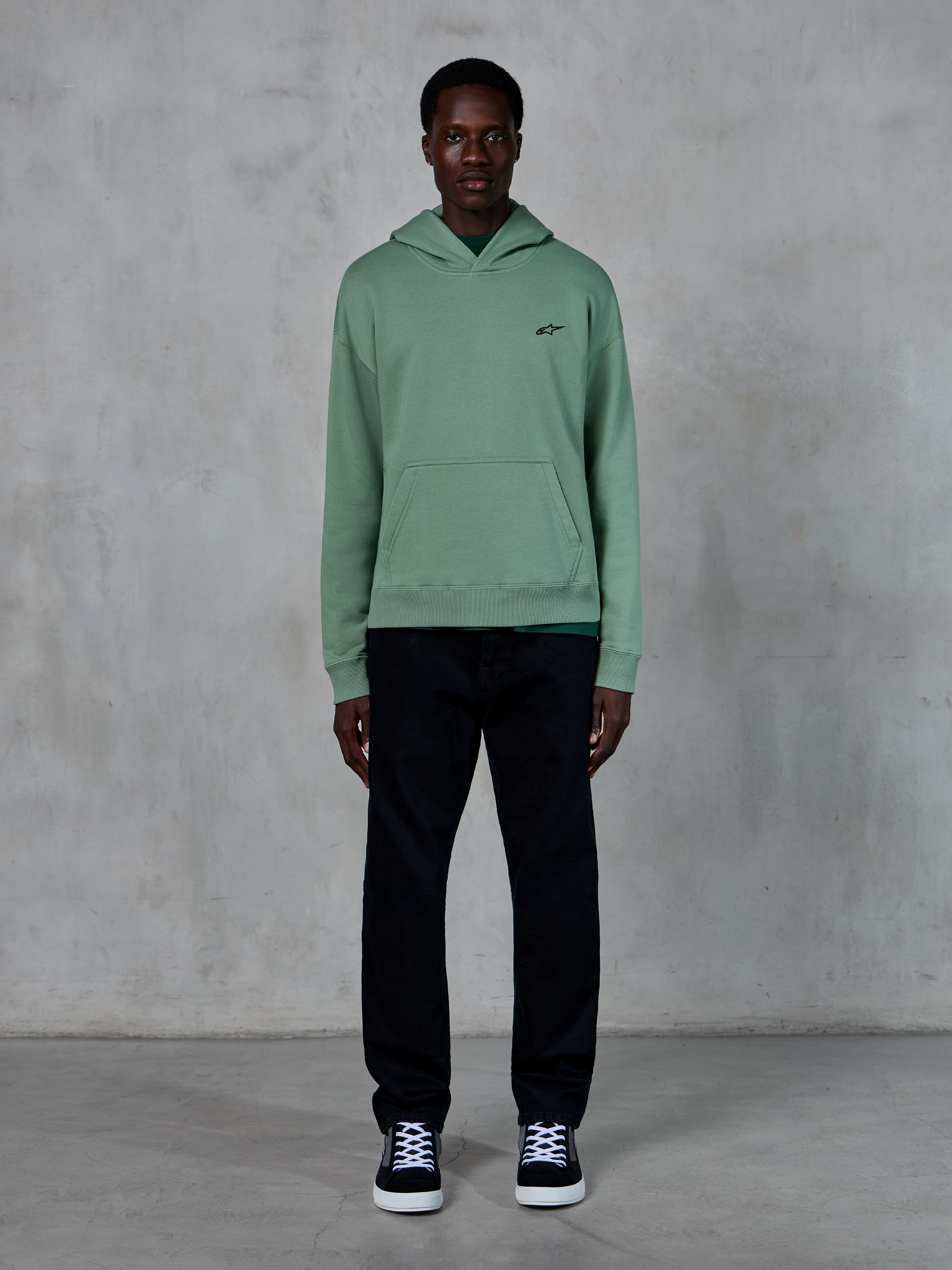 Alpinestars Essentials Ageless Hoodie, Cactus green, small black chest logo, kangaroo pocket, casual lifestyle streetwear worn with black pants and black and white sneakers against a textured wall