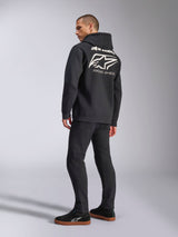 Modelo con la sudadera con capucha Formulation de Alpinestars, negra, vista trasera con un gran logotipo blanco de Alpinestars y la inscripción 'RACING AHEAD', estilo urbano inspirado en el motor con pantalones y zapatillas negras