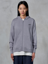 Alpinestars Essentials Ageless Zip Hoodie, color Silver, estilo urbano informal, sudadera de forro polar con cremallera completa y pequeño logo negro de Alpinestars en el pecho, bolsillo de canguro dividido, lucida por una modelo sobre una camiseta blanca y vaqueros oscuros