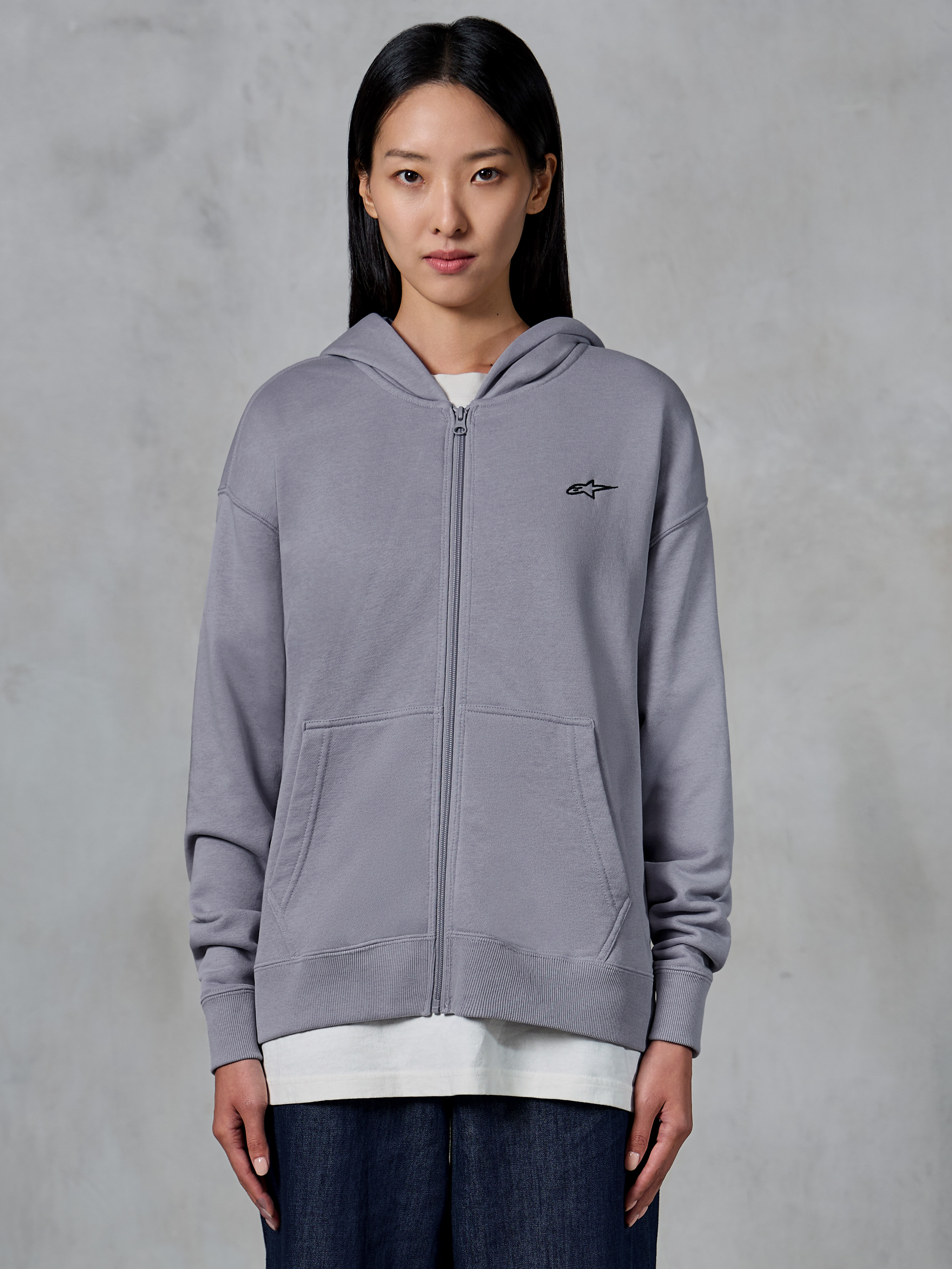 Alpinestars Essentials Ageless Zip Hoodie, color Silver, estilo urbano informal, sudadera de forro polar con cremallera completa y pequeño logo negro de Alpinestars en el pecho, bolsillo de canguro dividido, lucida por una modelo sobre una camiseta blanca y vaqueros oscuros