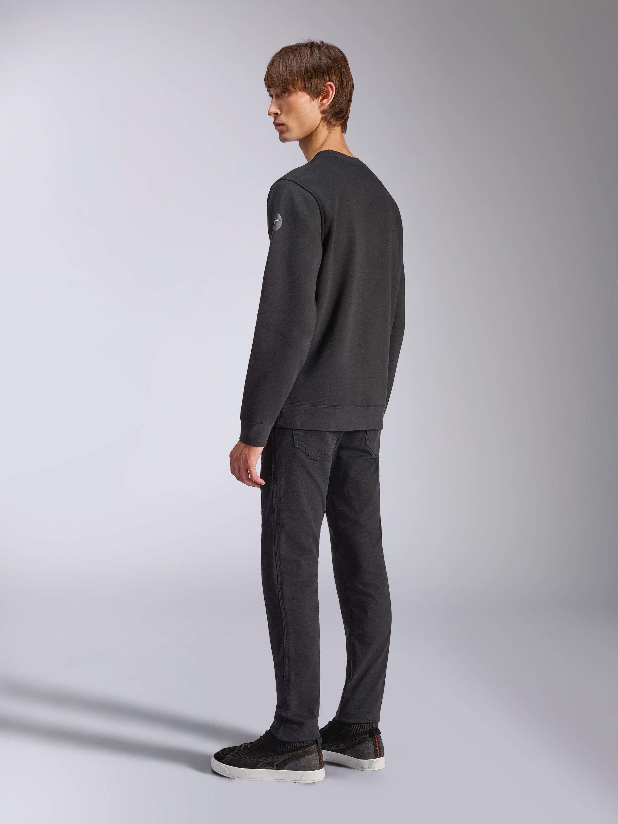 Alpinestars Bettern Crew, sudadera de forro polar negra, modelo masculino visto desde un ángulo lateral y posterior, con puños y dobladillo de canalé y un pequeño logotipo en la manga, estilo urbano casual inspirado en el automovilismo