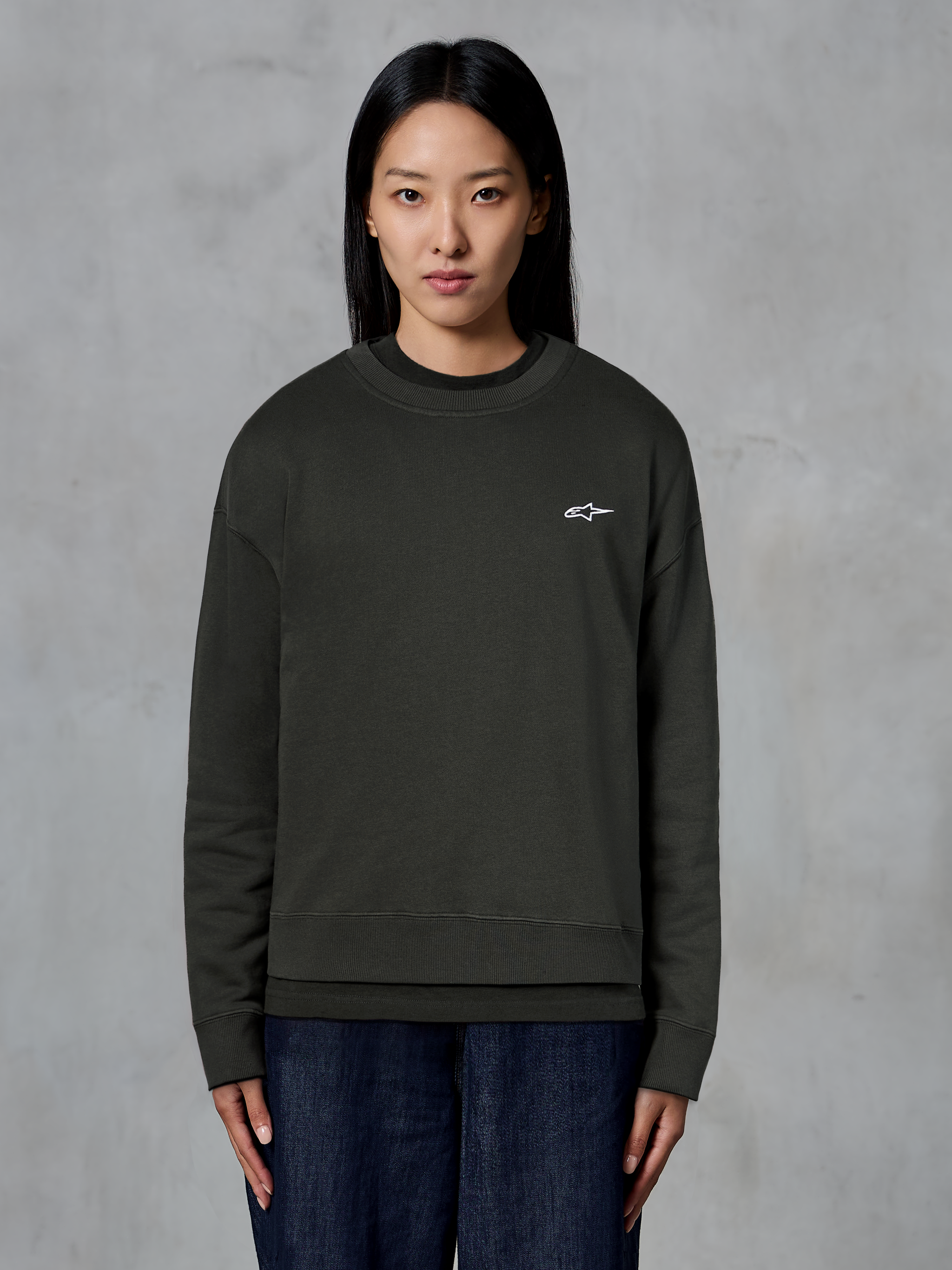 Alpinestars Essentials Ageless Crew, sudadera de cuello redondo premium, negro, con un pequeño logo blanco de Alpinestars en el pecho, estilo streetwear casual usada por una modelo con vaqueros oscuros