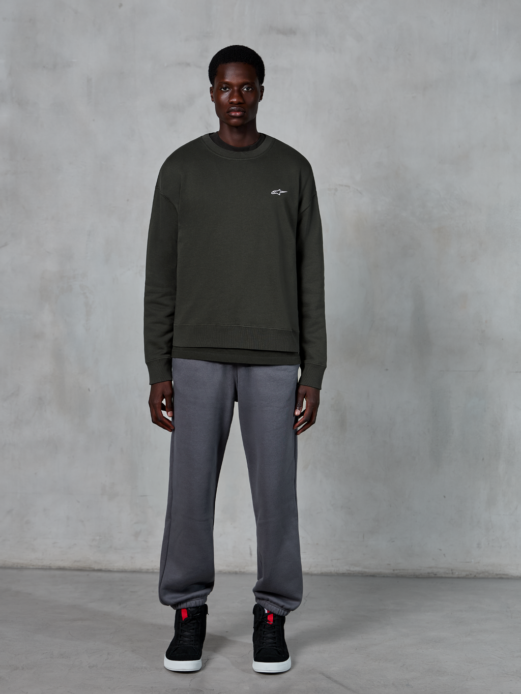 Alpinestars Essentials Ageless Crew, sudadera de cuello redondo verde oscuro con un pequeño logo blanco en el pecho, usada por un modelo con pantalones de chándal grises y zapatillas altas negras, ropa de estilo de vida casual