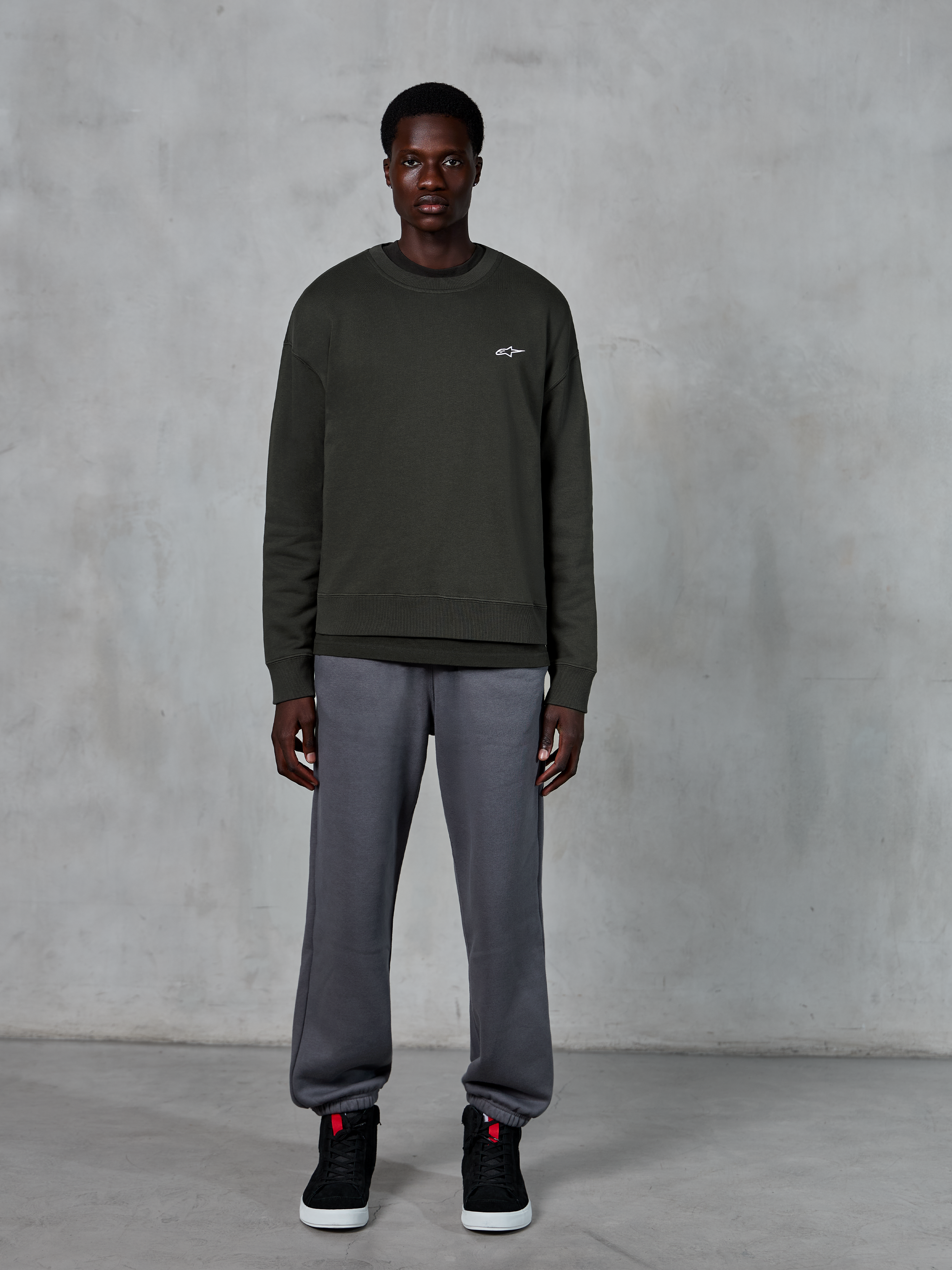 Alpinestars Essentials Ageless Crew, sudadera de cuello redondo verde oscuro con un pequeño logo blanco en el pecho, usada por un modelo con pantalones de chándal grises y zapatillas altas negras, ropa de estilo de vida casual