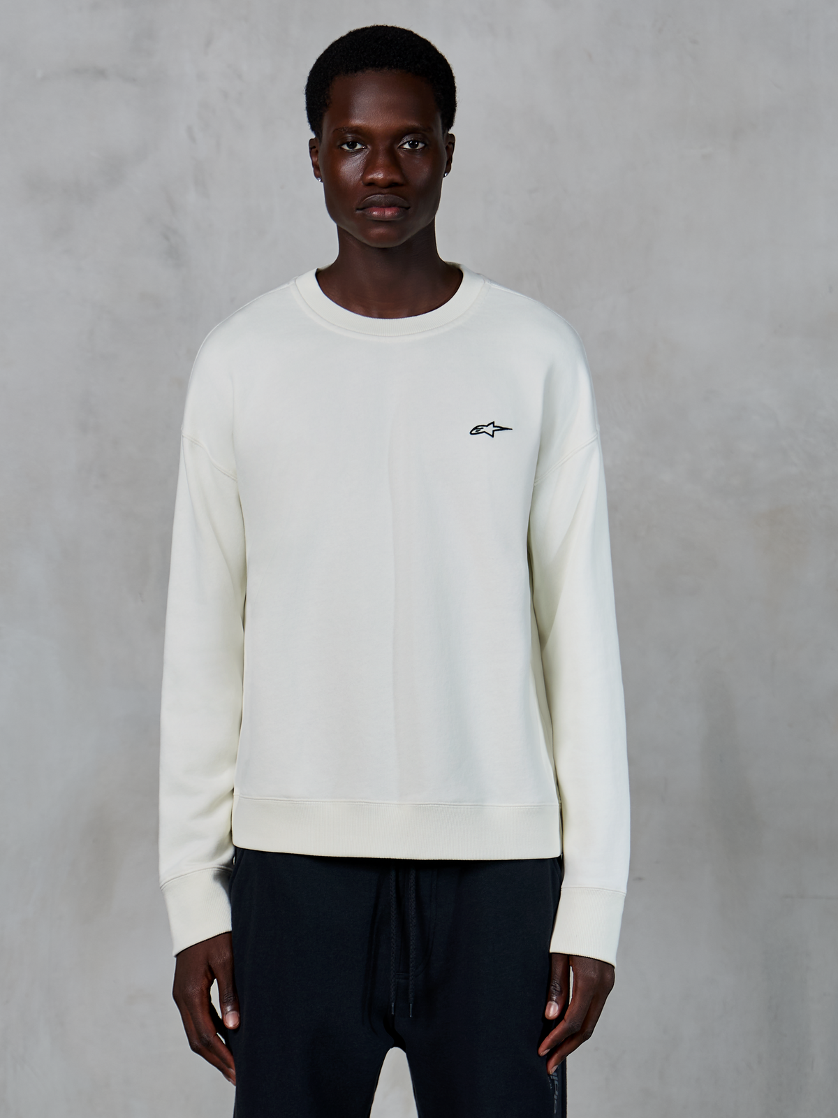 Alpinestars Essentials Ageless Crew, sudadera de cuello redondo en color blanco roto, pequeño logotipo negro de Astars en el pecho izquierdo, ropa deportiva de estilo casual, textura de forro polar premium, lucida por un modelo con pantalones joggers negros