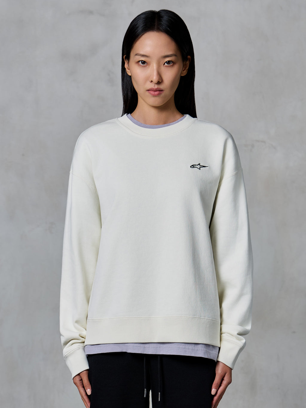Alpinestars Essentials Ageless Crew, sudadera de cuello redondo, blanco roto, con un pequeño logotipo de Alpinestars en negro en el pecho, ropa casual de estilo de vida modelada por una mujer