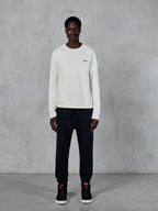 Alpinestars Essentials Ageless Crew, sudadera de cuello redondo, blanco roto con un pequeño logotipo de estrella negro, usada por un modelo con joggers negros, estilo streetwear casual inspirado en el automovilismo