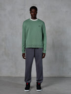 Alpinestars Essentials Ageless Crew, sudadera de cuello redondo en verde cactus, con un pequeño logotipo negro en el pecho, llevada por un modelo con pantalones de chándal grises y zapatillas negras, ropa urbana informal de inspiración motorsport