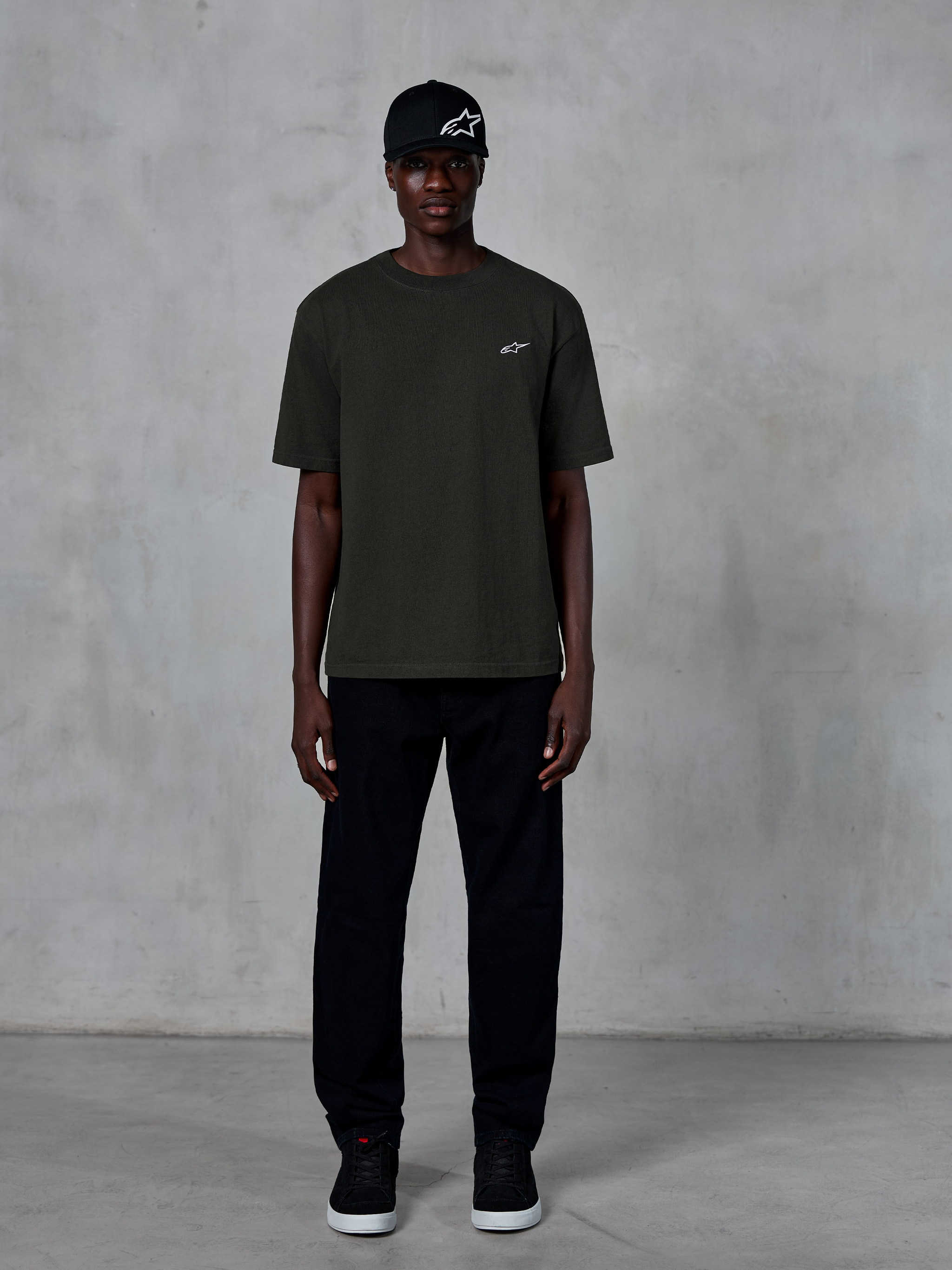 Camiseta Alpinestars Essentials Ageless SS, camiseta de manga corta, color negro, con un pequeño logotipo blanco de Alpinestars en el pecho, lucida por un modelo con gorra y pantalones negros para un look lifestyle impecable