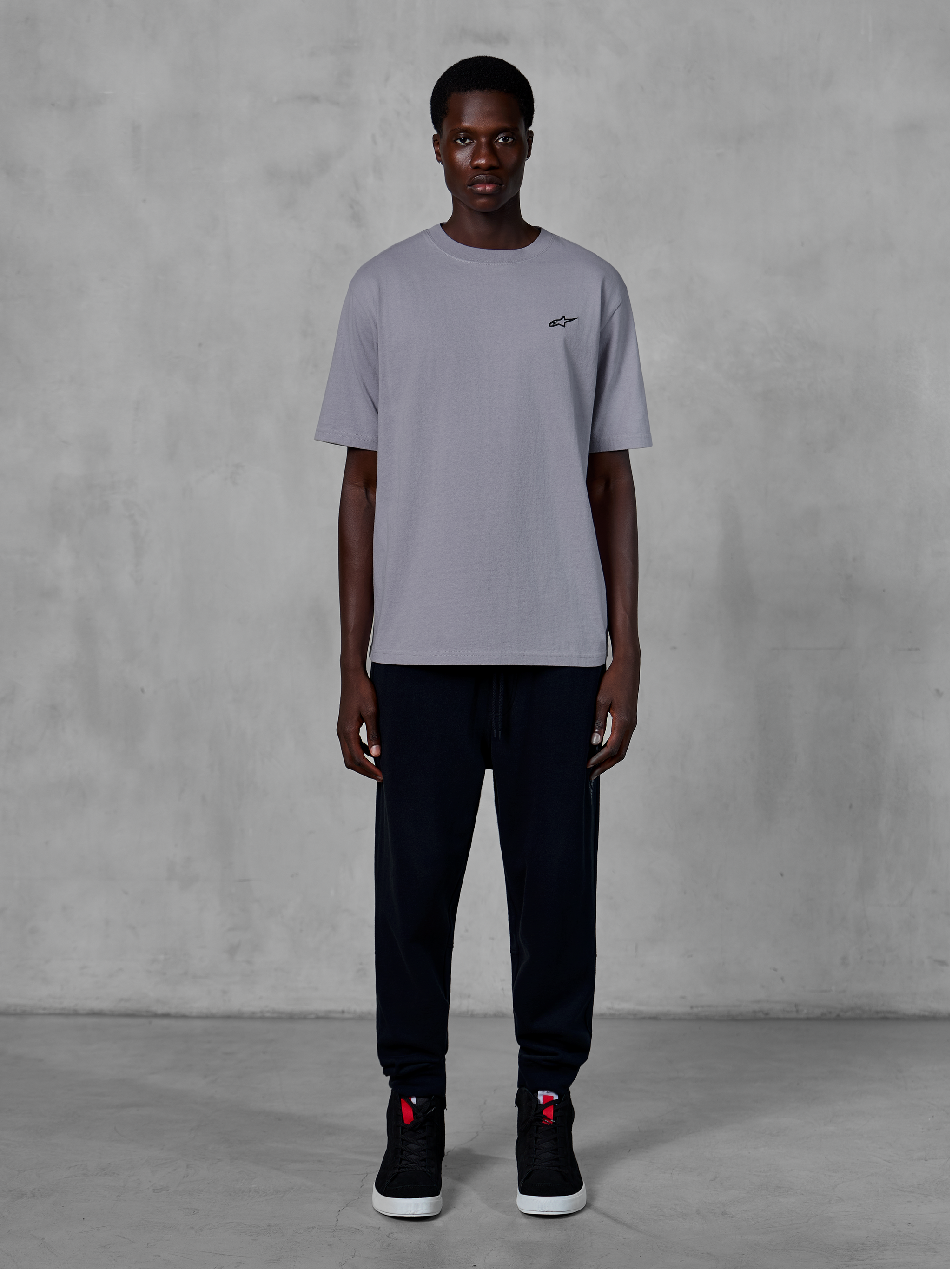 Alpinestars Essentials Ageless SS Tee, camiseta de manga corta, plateada, con un pequeño logotipo negro en el pecho, combinada con pantalones joggers y zapatillas negras, estilo streetwear casual de motorsport