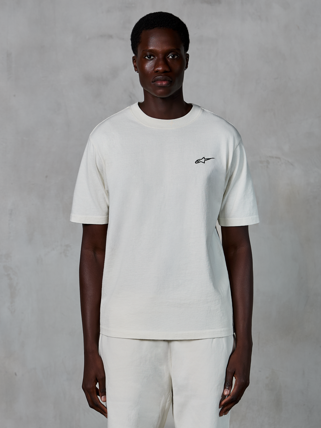 Camiseta Alpinestars Essentials Ageless SS, camiseta de manga corta, blanco roto, usada por un modelo masculino, con un pequeño logotipo negro de Alpinestars en el pecho, ajuste relajado, tejido de punto de algodón orgánico, estilo casual streetwear de motor
