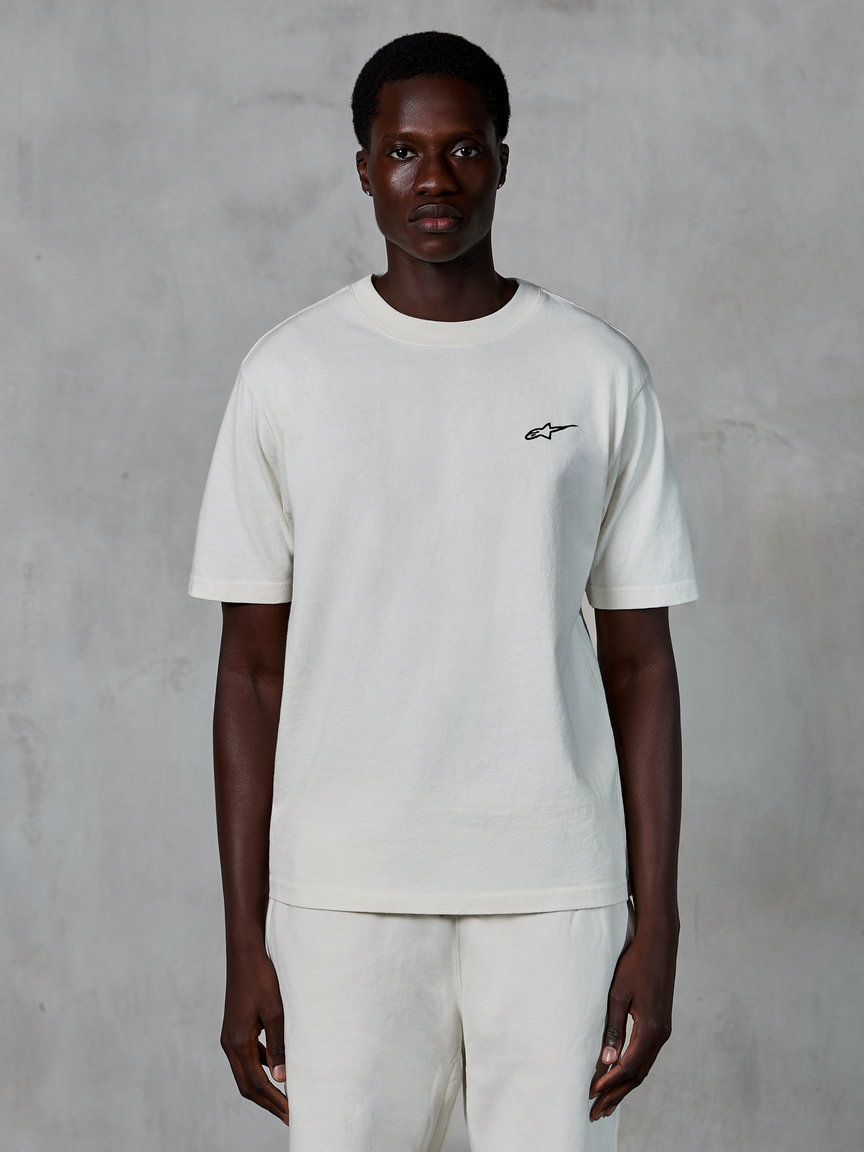 Camiseta Alpinestars Essentials Ageless SS, camiseta de manga corta, blanco roto, usada por un modelo masculino, con un pequeño logotipo negro de Alpinestars en el pecho, ajuste relajado, tejido de punto de algodón orgánico, estilo casual streetwear de motor