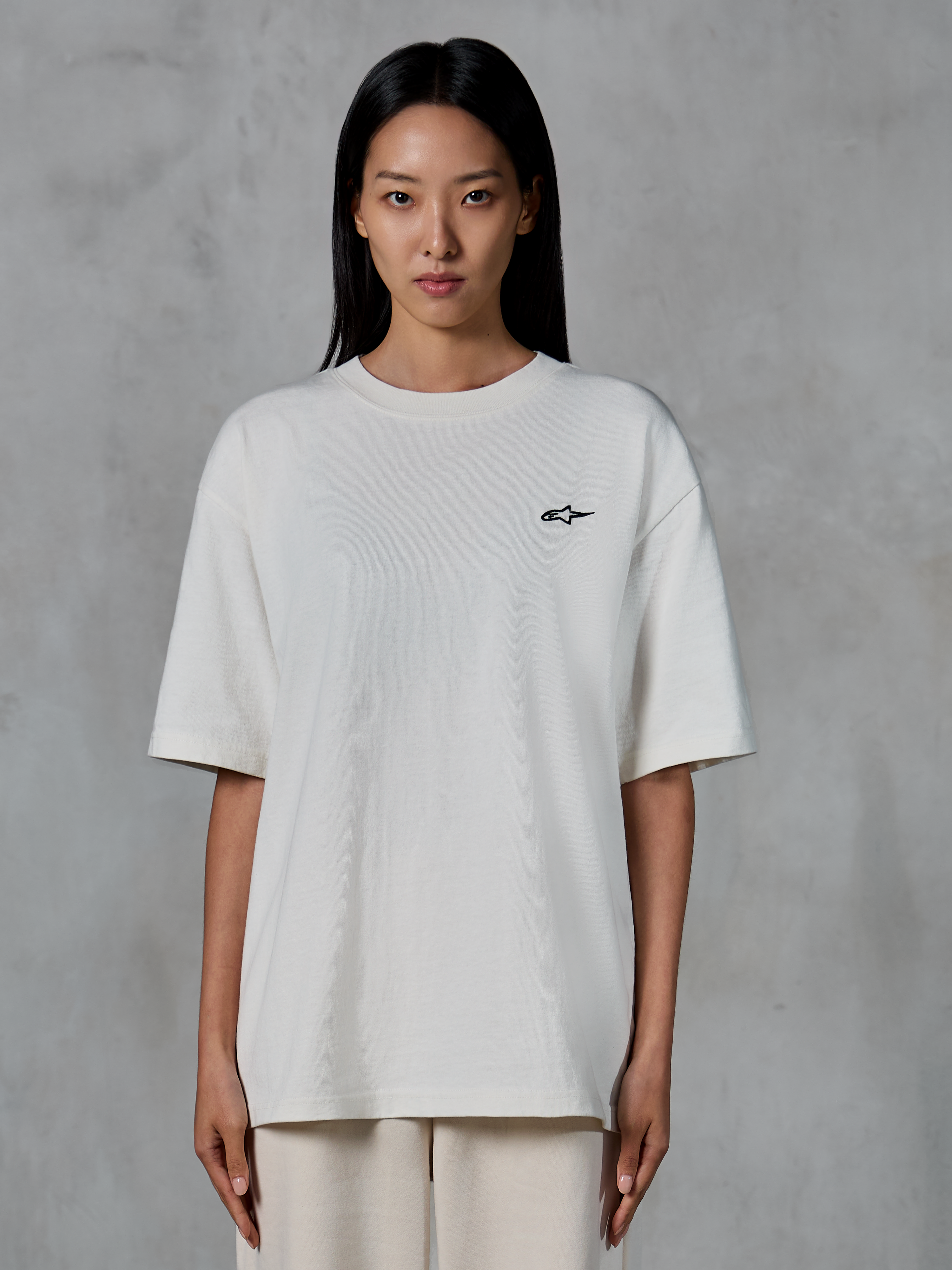 Camiseta Alpinestars Essentials Ageless SS, camiseta de manga corta, blanco roto, lucida por una modelo, con un pequeño logotipo negro de Alpinestars en el pecho, ajuste relajado, estilo de vida urbano casual