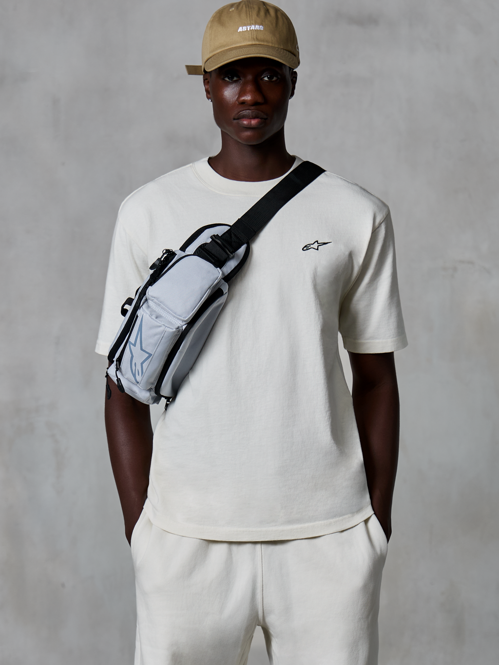 Alpinestars Essentials Ageless SS Tee, camiseta de manga corta, blanco roto, combinada con pantalones cortos a juego y una gorra color canela, con un pequeño logotipo negro en el pecho y un bolso bandolera gris, estilo urbano informal inspirado en el motorsport