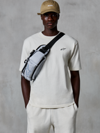 Alpinestars Essentials Ageless SS Tee, camiseta de manga corta, blanco roto, combinada con pantalones cortos a juego y una gorra color canela, con un pequeño logotipo negro en el pecho y un bolso bandolera gris, estilo urbano informal inspirado en el motorsport