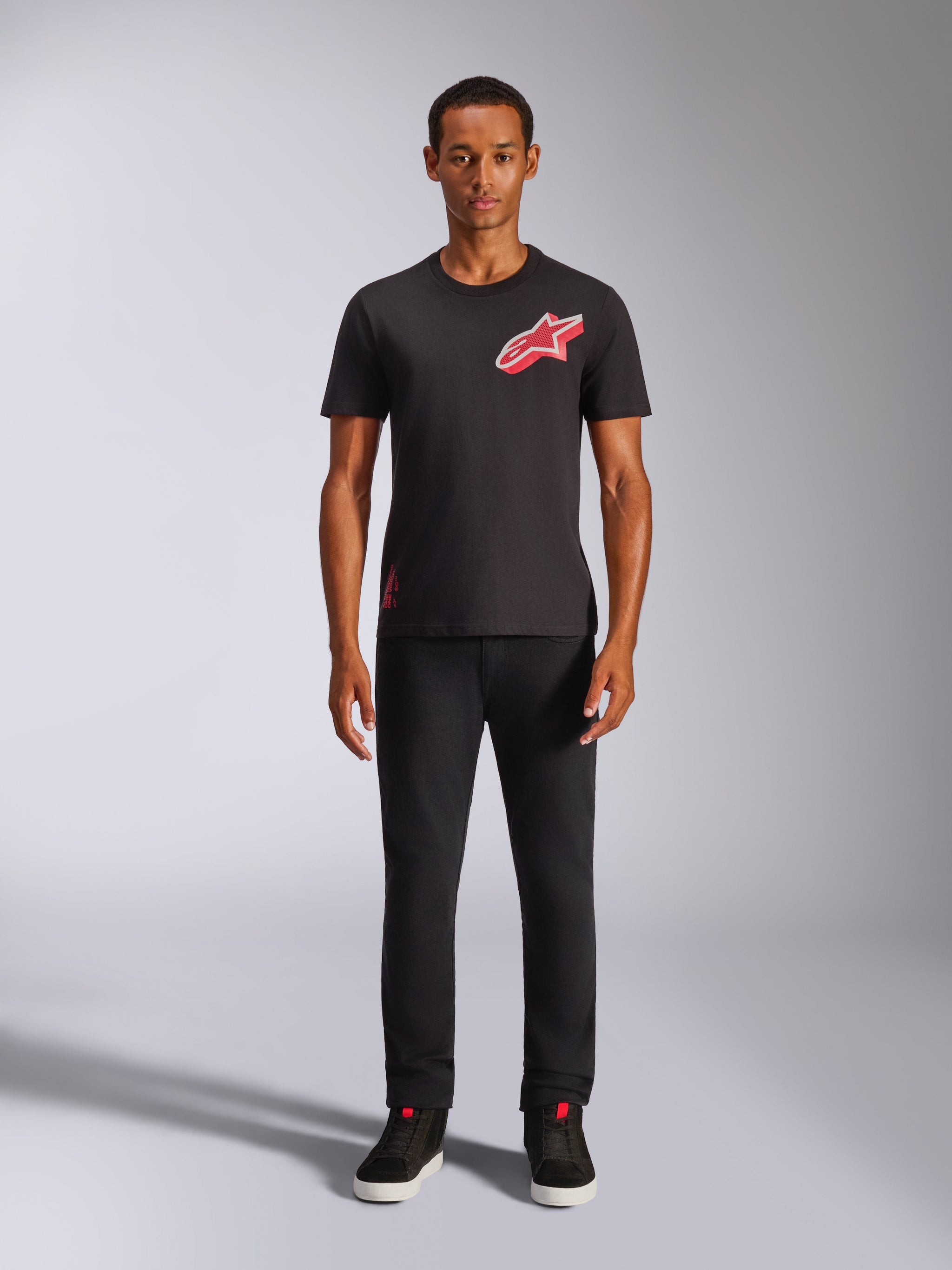 Modelo masculino luciendo la camiseta Alpinestars Shaded CSF de manga corta en color negro, con un logotipo Astars en lámina con degradado rojo inclinado en el pecho, combinado con pantalones negros y zapatillas, estilo streetwear casual de automovilismo