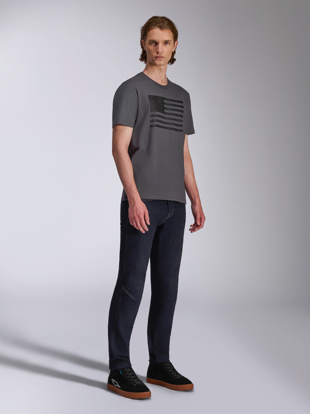Alpinestars Waving CSF Tee, camiseta de manga corta, gris carbón, gráfico de bandera ondeante negra, usada por un modelo con vaqueros oscuros y zapatillas, ropa urbana casual de estilo motorsport