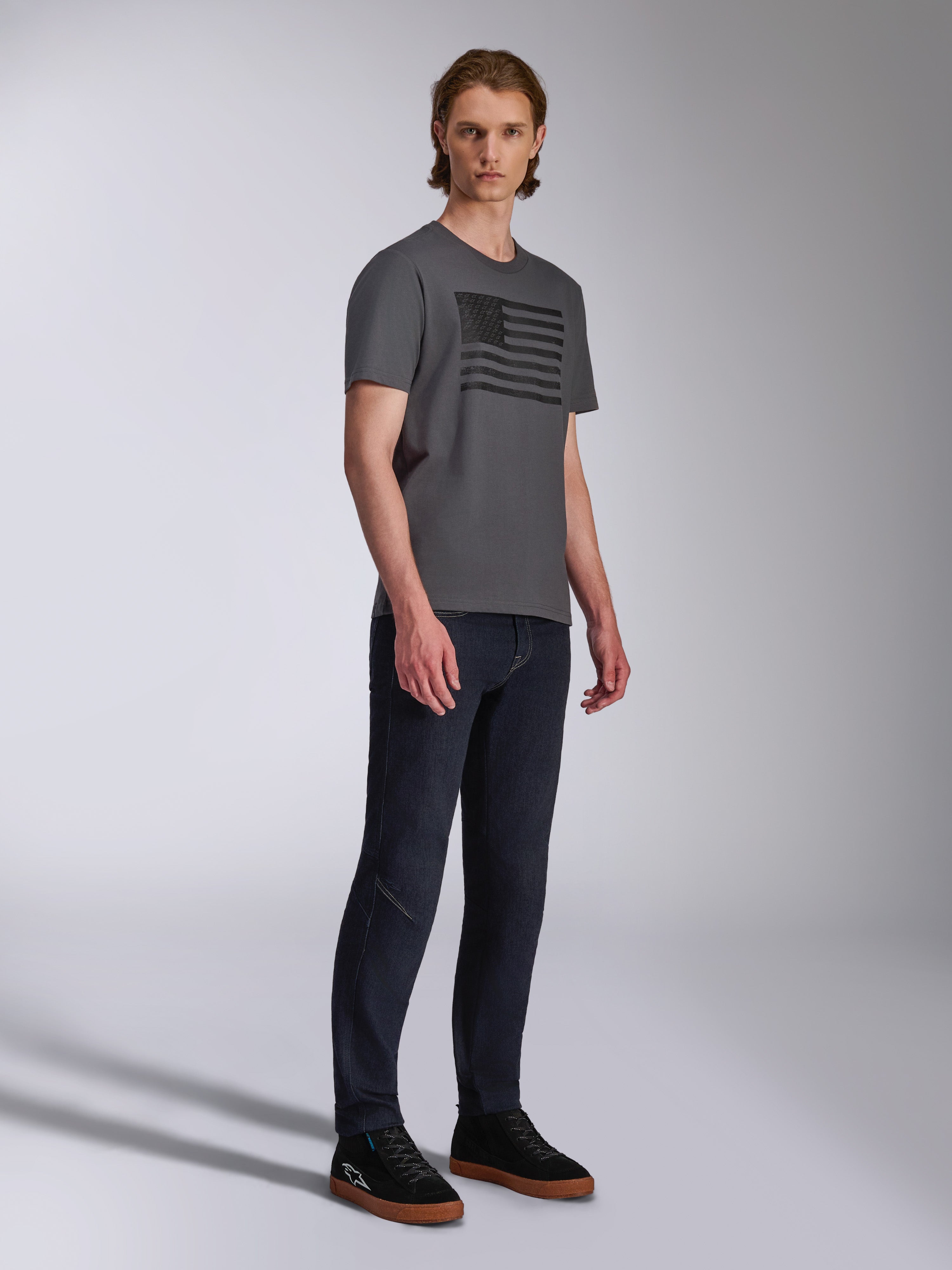 Alpinestars Waving CSF Tee, camiseta de manga corta, gris carbón, gráfico de bandera ondeante negra, usada por un modelo con vaqueros oscuros y zapatillas, ropa urbana casual de estilo motorsport