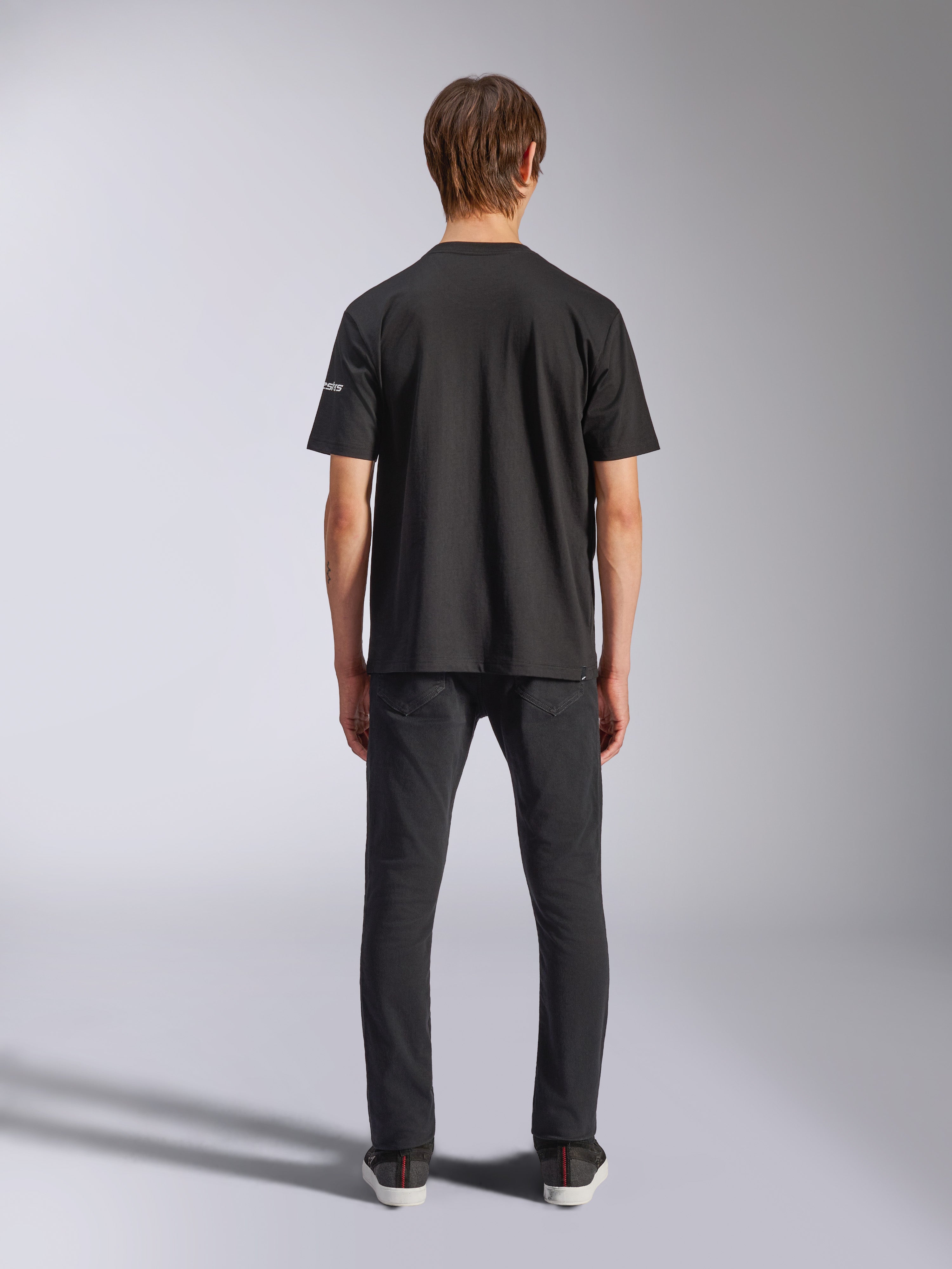 Alpinestars Frontal CSF Tee, camiseta de manga corta, negra, vista posterior en modelo con vaqueros negros, ropa de calle casual de alto rendimiento con detalle de logotipo blanco pequeño en la manga