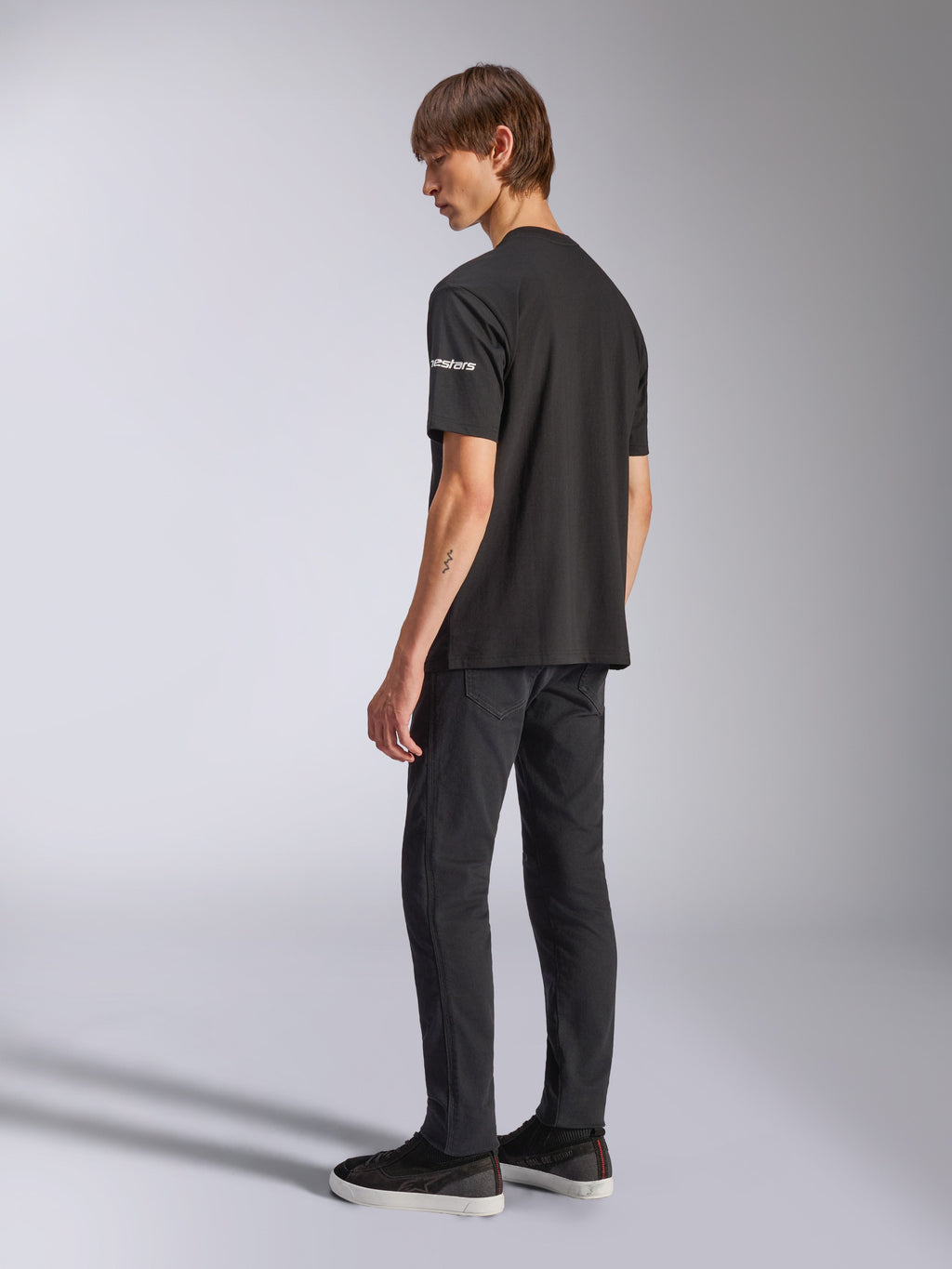 Camiseta Alpinestars Frontal CSF, camiseta de manga corta, negra, logotipo astars blanco en la manga, usada por un modelo con pantalones y zapatos negros, diseño casual inspirado en el automovilismo