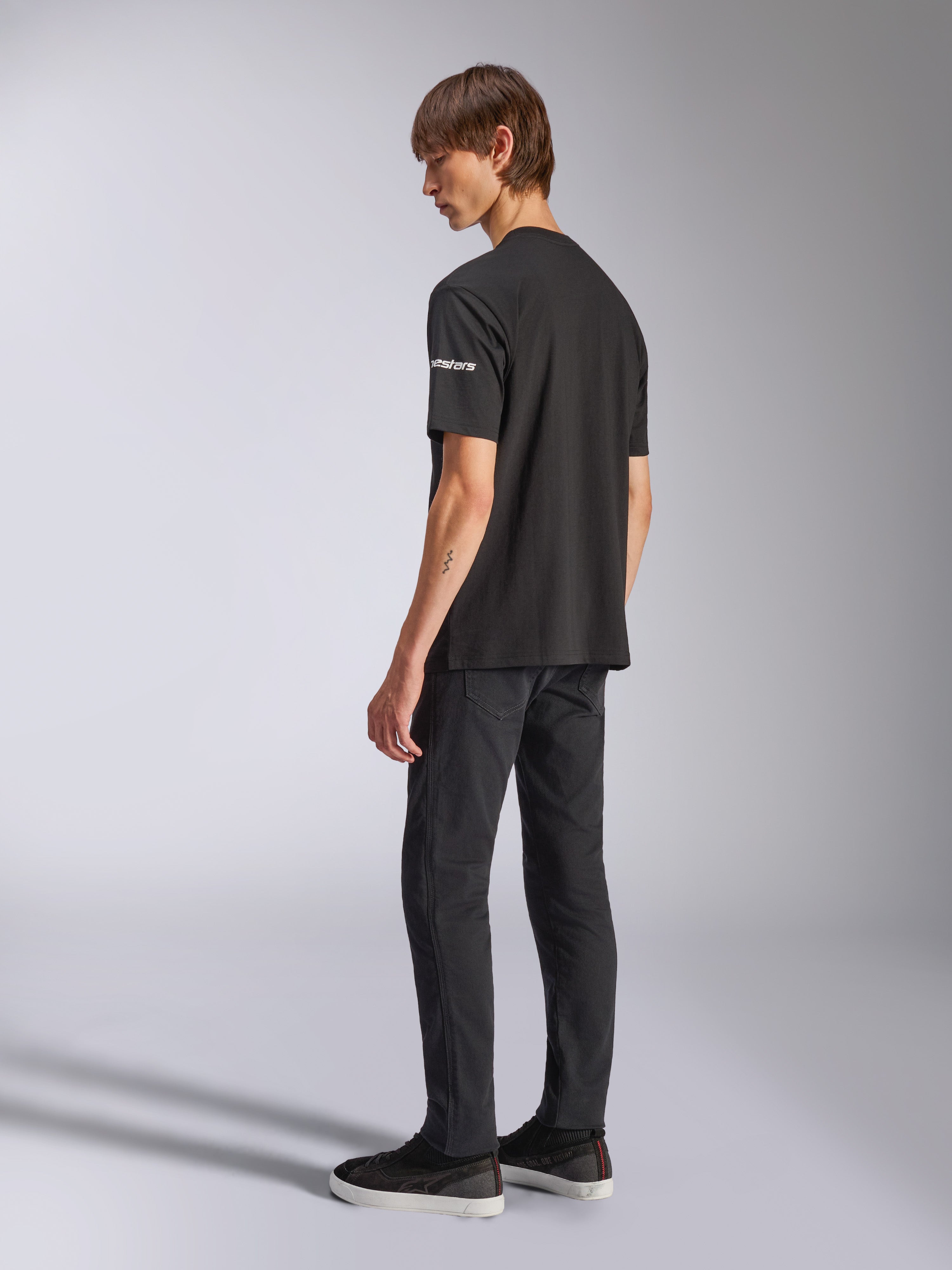 Camiseta Alpinestars Frontal CSF, camiseta de manga corta, negra, logotipo astars blanco en la manga, usada por un modelo con pantalones y zapatos negros, diseño casual inspirado en el automovilismo