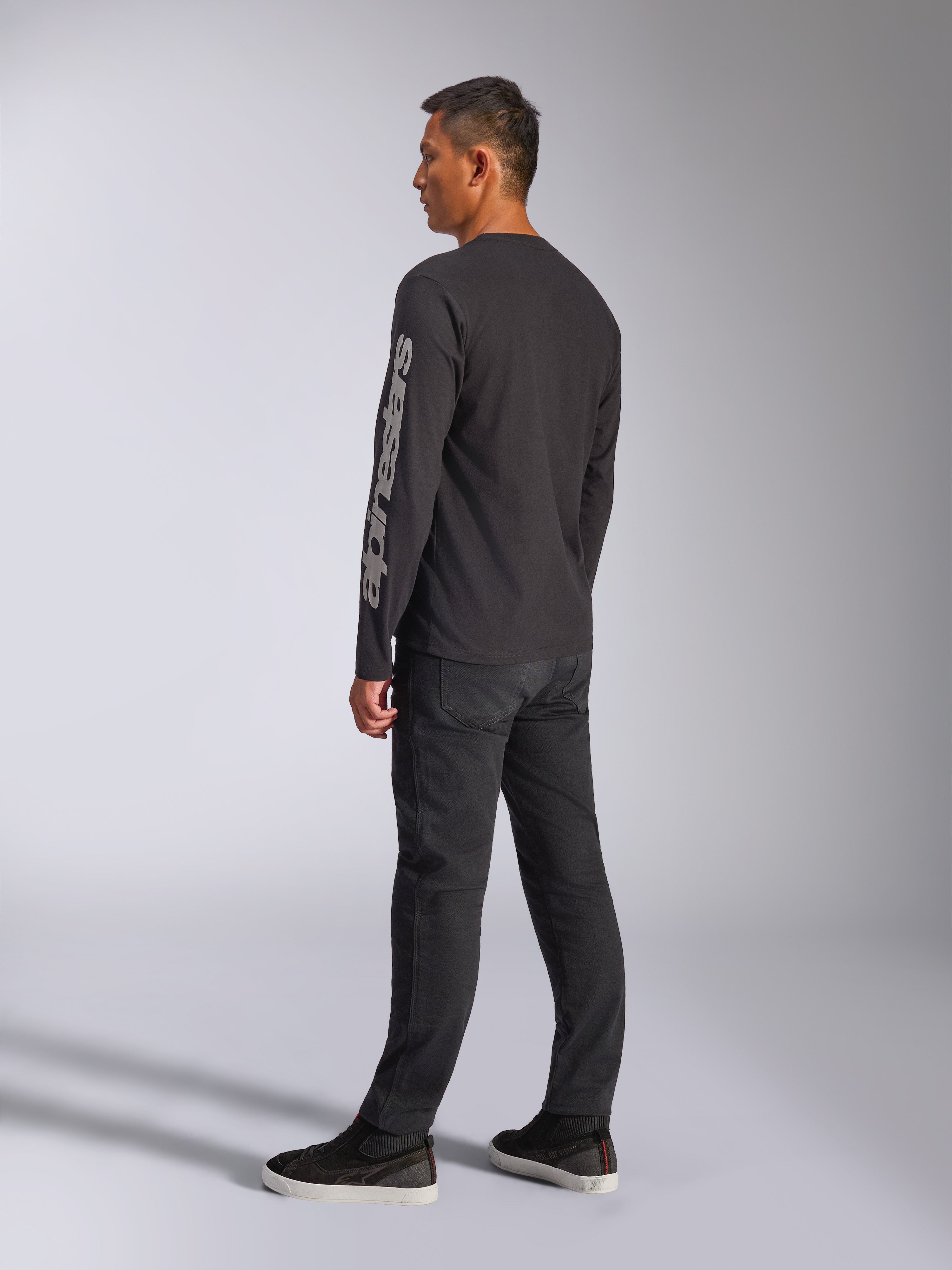 Camiseta de manga larga Alpinestars Trackside CSF, negra, con un gran logotipo gris de Alpinestars en la manga izquierda, lucida por un modelo con pantalones negros y zapatillas casuales, estilo streetwear casual inspirado en el automovilismo.