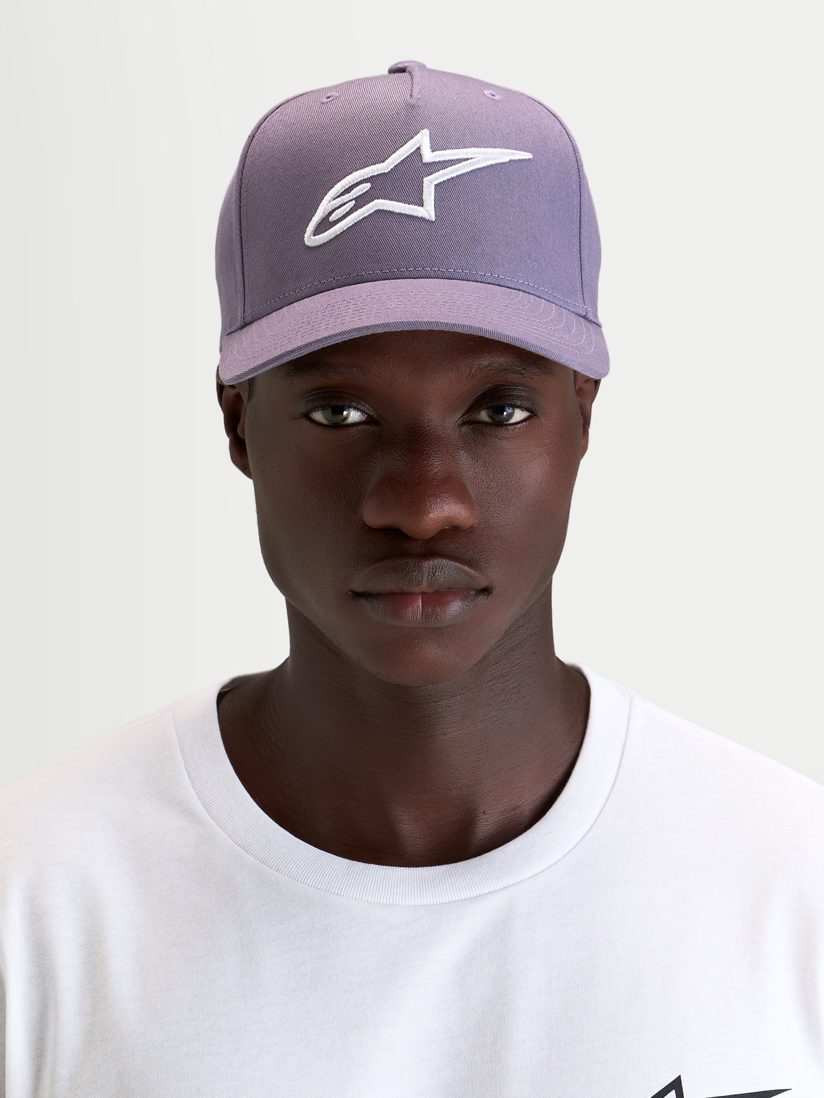 Gorra Ageless Snapback Alpinestars, gorra deportiva con visera curva, morada y blanca, con el logo Astars bordado en 3D en el panel frontal, vista frontal en modelo