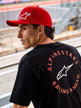 Gorra Alpinestars Corp Shift Multi, gorra roja de visera curva con logo Astars en 3D blanco, combinada con una camiseta Alpinestars negra con el gráfico 'Racing Ahead', en un entorno de motor lifestyle en un circuito de carreras