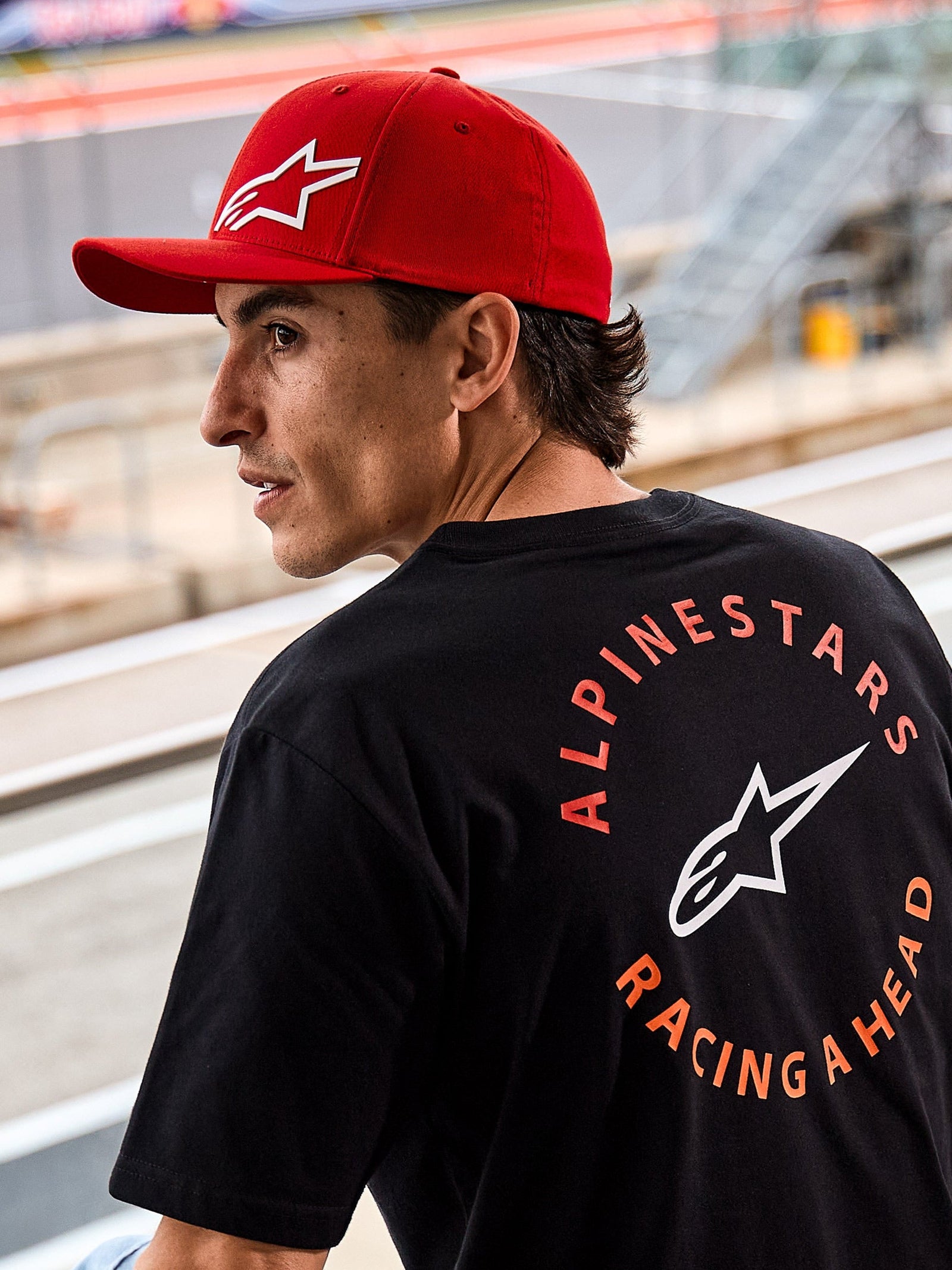 Gorra Alpinestars Corp Shift Multi, gorra roja de visera curva con logo Astars en 3D blanco, combinada con una camiseta Alpinestars negra con el gráfico 'Racing Ahead', en un entorno de motor lifestyle en un circuito de carreras
