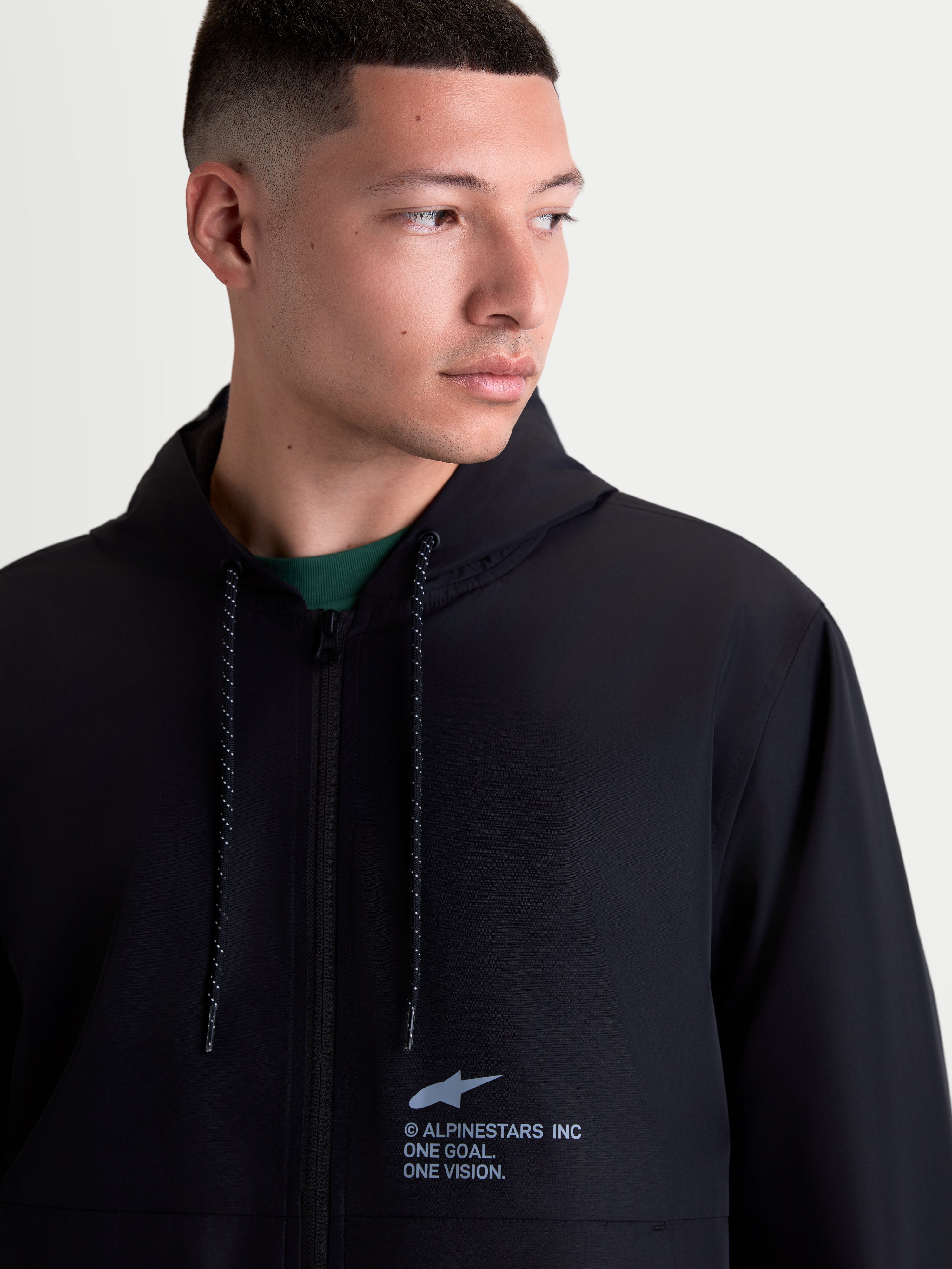 Alpinestars Chaqueta Cortavientos Speeder Resolve, cortavientos ligero con capucha para hombre, negro, de poliéster, vista frontal en modelo con logotipo en el pecho y cordones moteados.