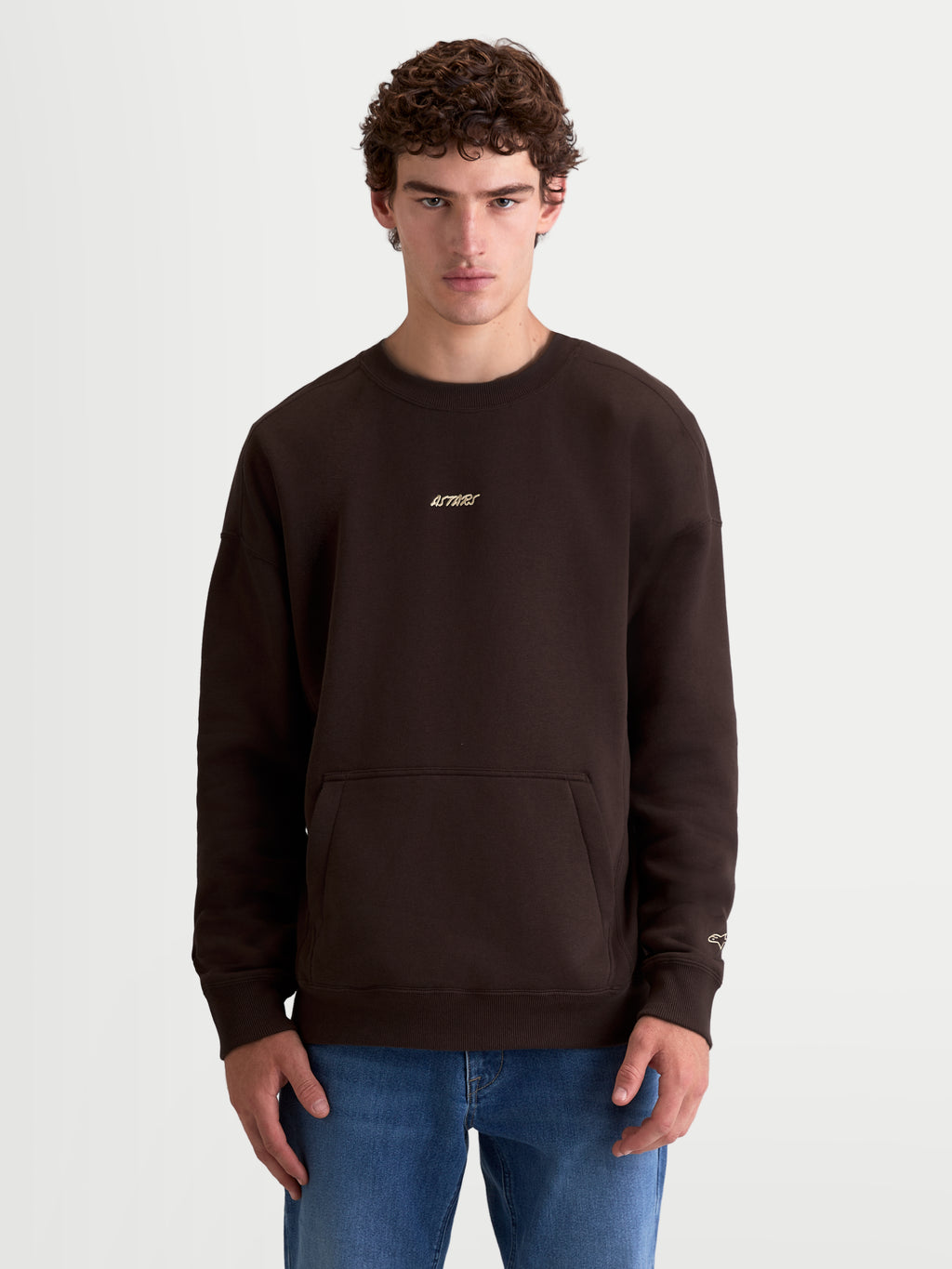 Alpinestars Guise Oversize, sudadera de forro polar con cuello redondo, ajuste relajado unisex mostrado en modelo, color marrón, confeccionada en algodón y poliéster reciclado, con bolsillo canguro y logotipos bordados en el pecho y la manga, estilo casual inspirado en el motorsport.