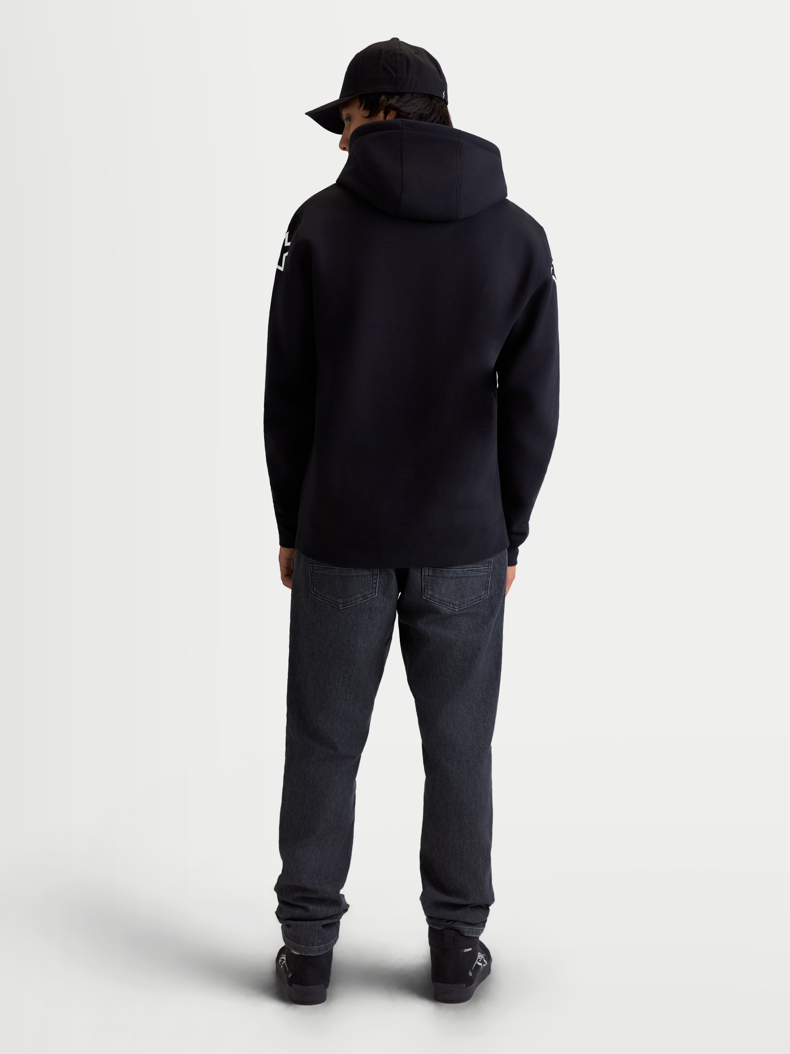 Alpinestars Sudadera Con Capucha Velocifier, ropa deportiva para hombre de forro polar, negra con logotipos blancos en los hombros, vista trasera en modelo que muestra la capucha, confeccionada en mezcla de algodón y poliéster reciclado.