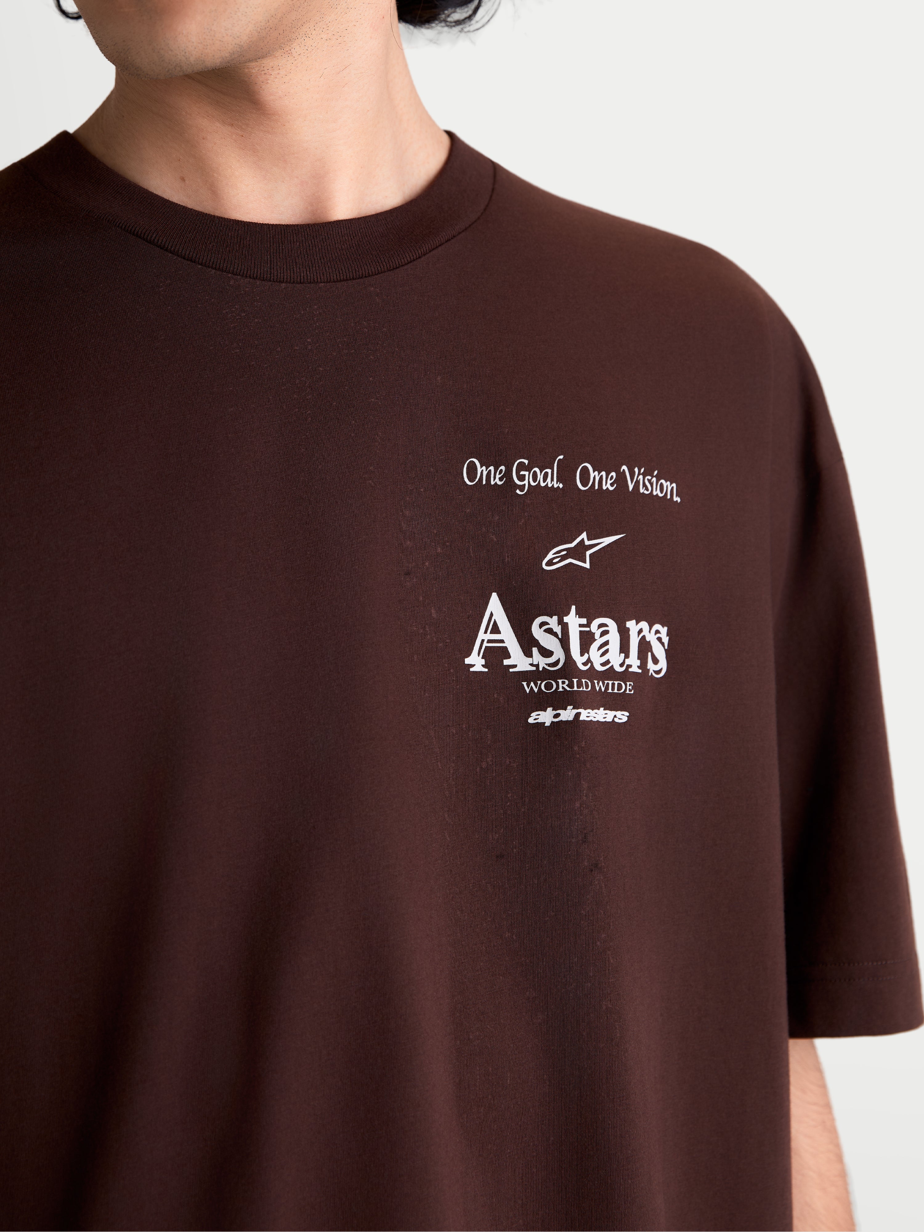 Alpinestars Camiseta Oversized Post CSF, camiseta de mangas cortas marrón, primer plano del gráfico blanco en el pecho con el lema One Goal. One Vision. y logotipo Astars worldwide, mezcla de algodón y poliéster