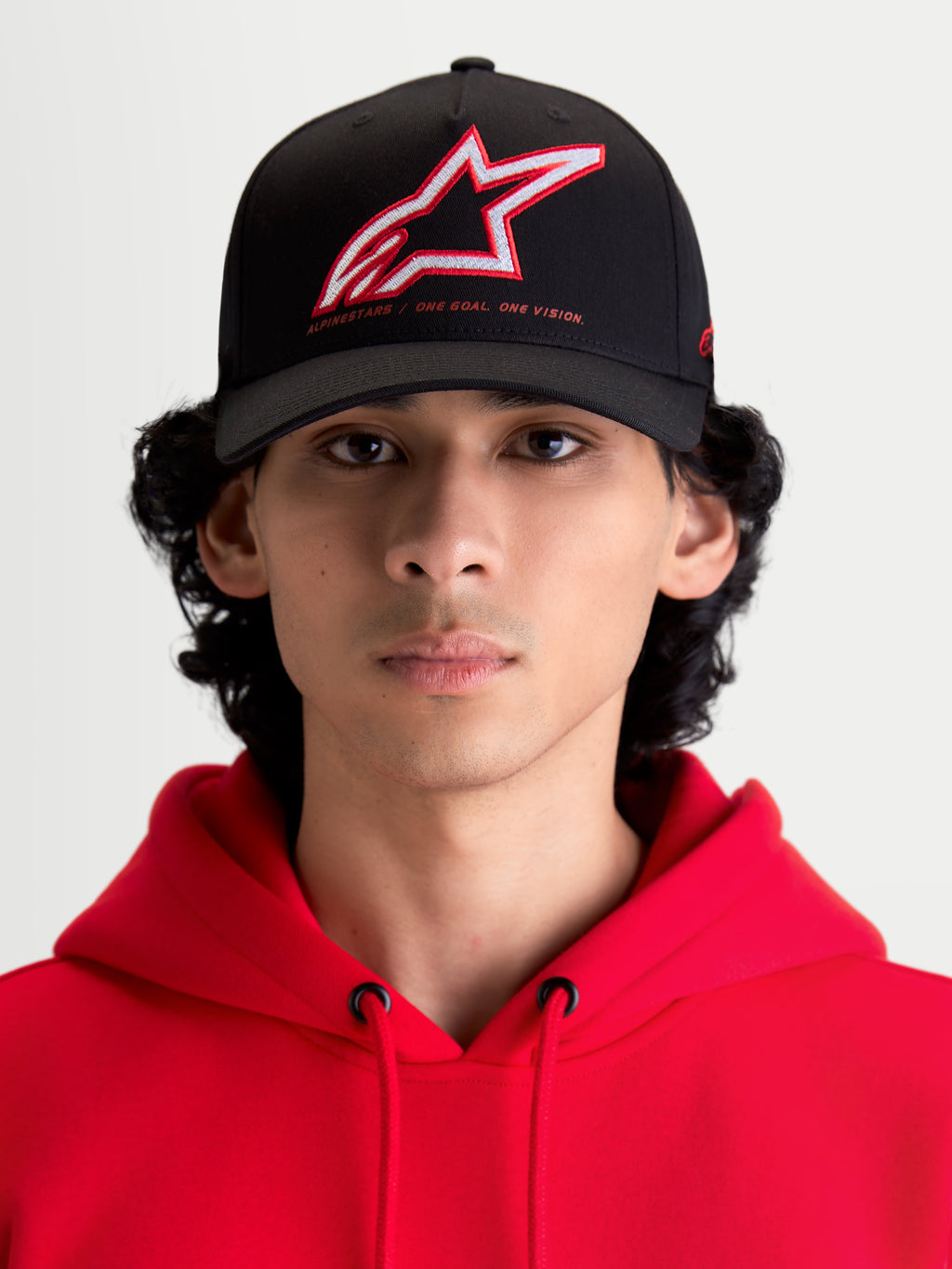Alpinestars Lineout, gorra de béisbol para sportswear con visera curva, negra con logotipo Astars bordado en rojo y blanco y lema One Goal One Vision, vista frontal en modelo con sudadera roja, construcción de sarga estructurada.