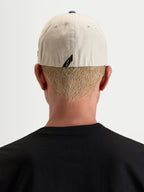 Gorra County Canvas Alpinestars, gorra sportswear casual, color arena y azul, vista trasera en modelo mostrando el ajuste flex-back y el detalle sutil de la etiqueta trasera