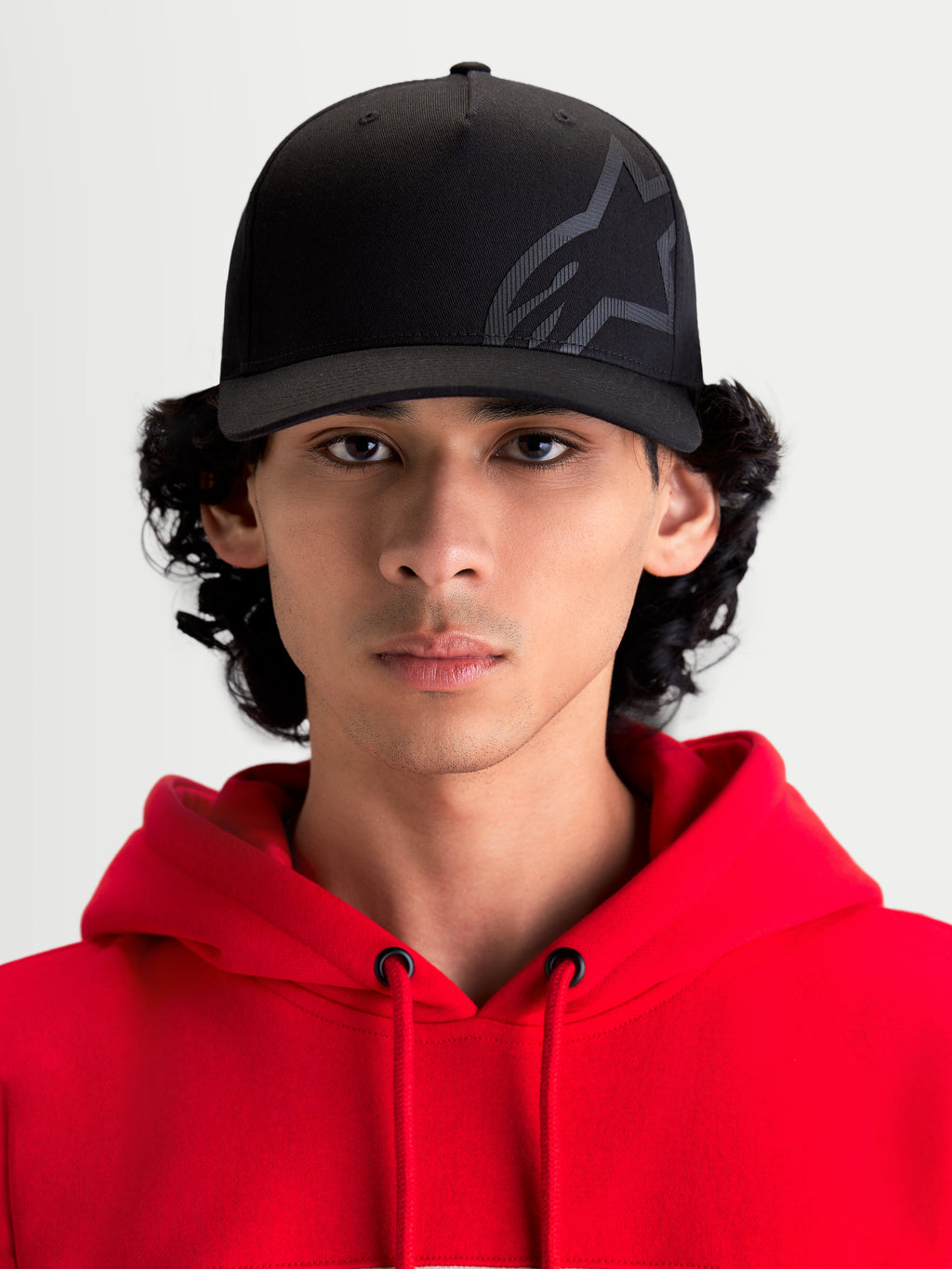Alpinestars Gorra Maze, gorra de estilo motorsport negra para uso casual, vista frontal en modelo con sudadera roja, corona estructurada, visera curva y logotipo Astars Corp Shift texturizado en tejido de sarga resistente para un estilo fuera de la moto